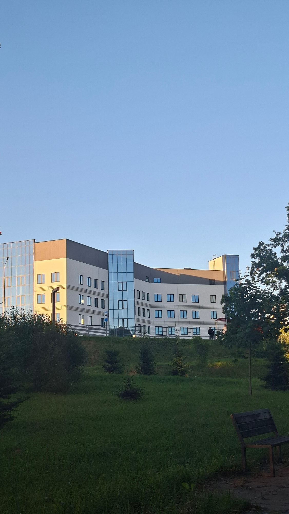 The image depicts a modern, multi-story building with a light-colored facade and dark gray accents, featuring prominent glass sections. The building appears to be located in an urban or suburban environment, with a clear blue sky overhead. In the foreground, there is a grassy area with several small evergreen trees and a wooden park bench. A larger tree with red berries is visible to the right of the building. The lighting suggests it is either early morning or late afternoon, with the sun casting long shadows. Based on the context provided, this scene is located in the city of Smalyavichy, Belarus. There are at least two figures visible near the building, one standing and another further back.