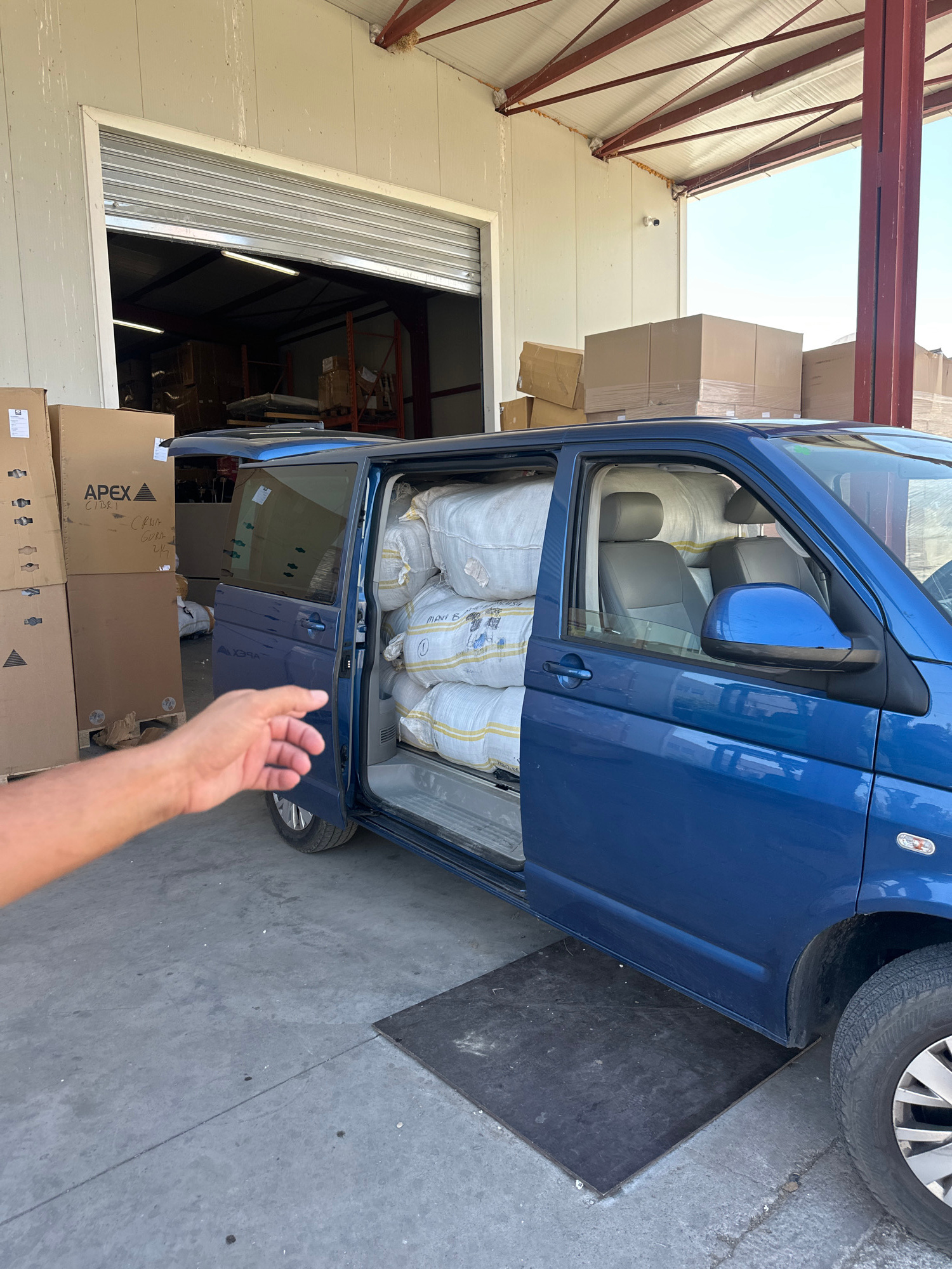 In an outdoor warehouse setting with a clear sky, a blue van is parked with its side door and rear doors open, revealing large white sacks filling the cargo area. The van is positioned in front of a large industrial building with corrugated metal walls and a metal beam structure overhead. Stacks of cardboard boxes are visible both inside the building and in front of the van.
A person's hand is reaching out into the frame from the lower right corner. The scene suggests a loading or unloading activity, with the open van and nearby boxes. The lighting indicates it is daytime. Text is visible on some of the boxes, including "APEX" and what appears to be product information. The location is identified as Rakića Kuće, Montenegro.