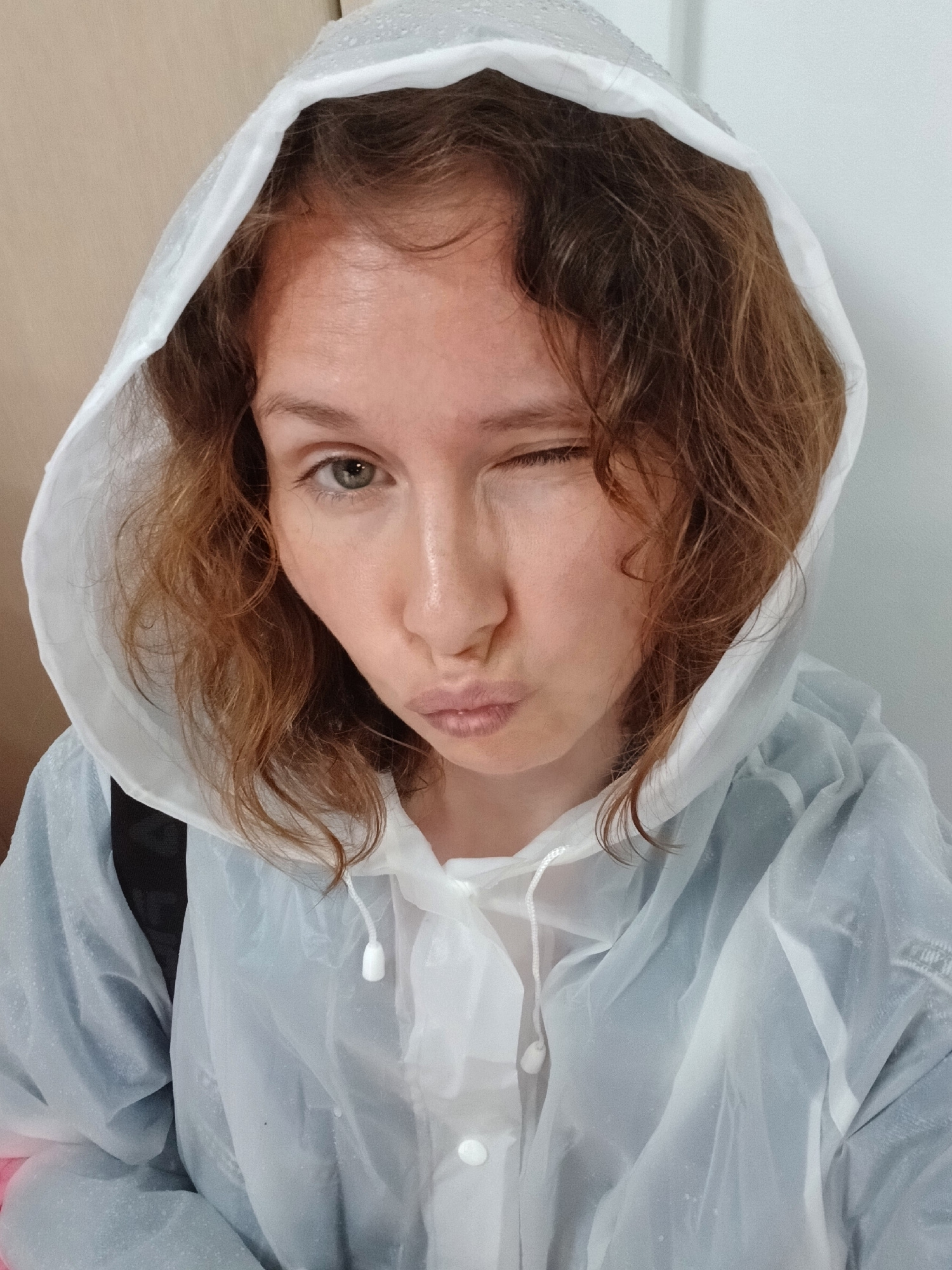 A young woman with curly, reddish-brown hair, wearing a transparent white hooded rain jacket, is depicted in a close-up selfie. She is winking with her left eye and puckering her lips. Her right eye is open, revealing a green iris. The background appears to be an indoor setting with a light-colored wall and a wooden-grained surface on the left. The lighting is even and diffused, suggesting an overcast or indoor environment. The image does not contain any information that would allow for determining the location as Yekaterinburg, Russia.