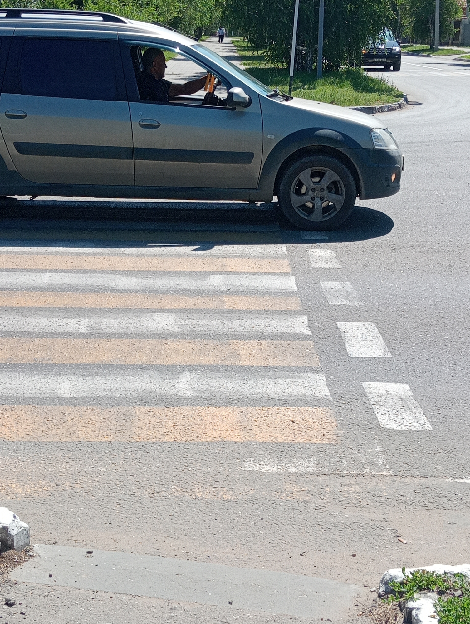 A side-view, medium shot captures a man driving a light-colored station wagon on an asphalt road. The man, with a shaved head and dark shirt, is visible through the car window, holding the steering wheel. The car is positioned on the right side of a pedestrian crossing marked with white and orange stripes. In the background, a second dark-colored SUV is stopped at a curve in the road, and beyond that, trees and buildings line the street. The scene appears to be in an urban environment, under bright daylight.