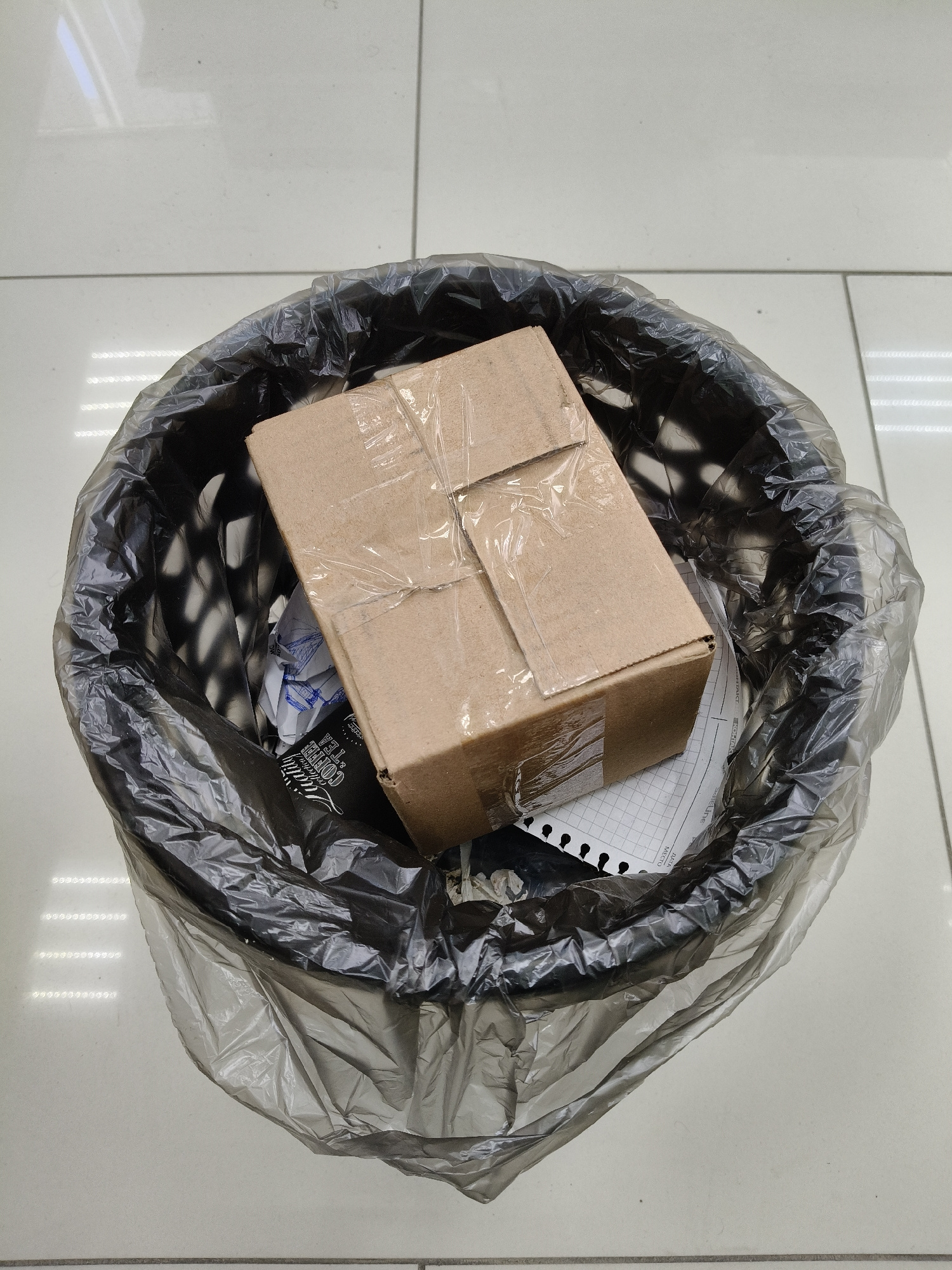 A cardboard box is placed in a black trash can lined with a translucent plastic bag, on a tiled floor. The box is taped shut with clear packing tape. Beneath the box, a crumpled piece of paper with grid lines is visible, along with other torn paper scraps. The trash can has a perforated pattern around its upper edge. The scene appears to be indoors, possibly in an office or a public space, given the tiled flooring.