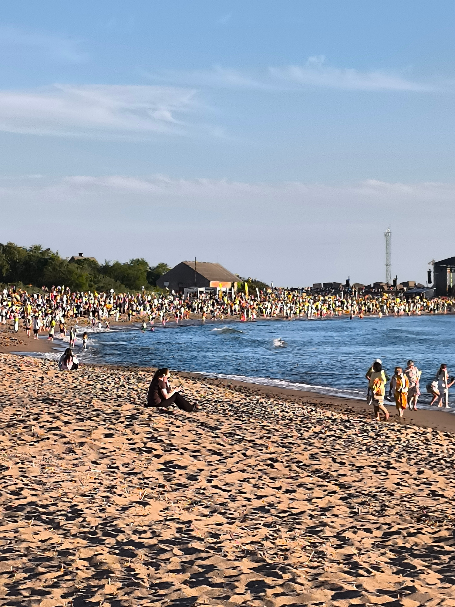 The image depicts a crowded beach scene, likely in Klaipėda, Lithuania, given the presence of a prominent radio tower in the background often associated with the city's coastline. The foreground is dominated by sand dunes and beachgoers, with two individuals sitting in the sand. The middle ground features a large number of people gathered along the shoreline and in the shallow water, suggesting a lively event or festival. In the background, there are buildings, including what appear to be tents or temporary structures, and the aforementioned radio tower. The sea is visible with gentle waves, and the sky is clear with scattered clouds. The overall impression is of a sunny day with a significant gathering of people enjoying the beach.