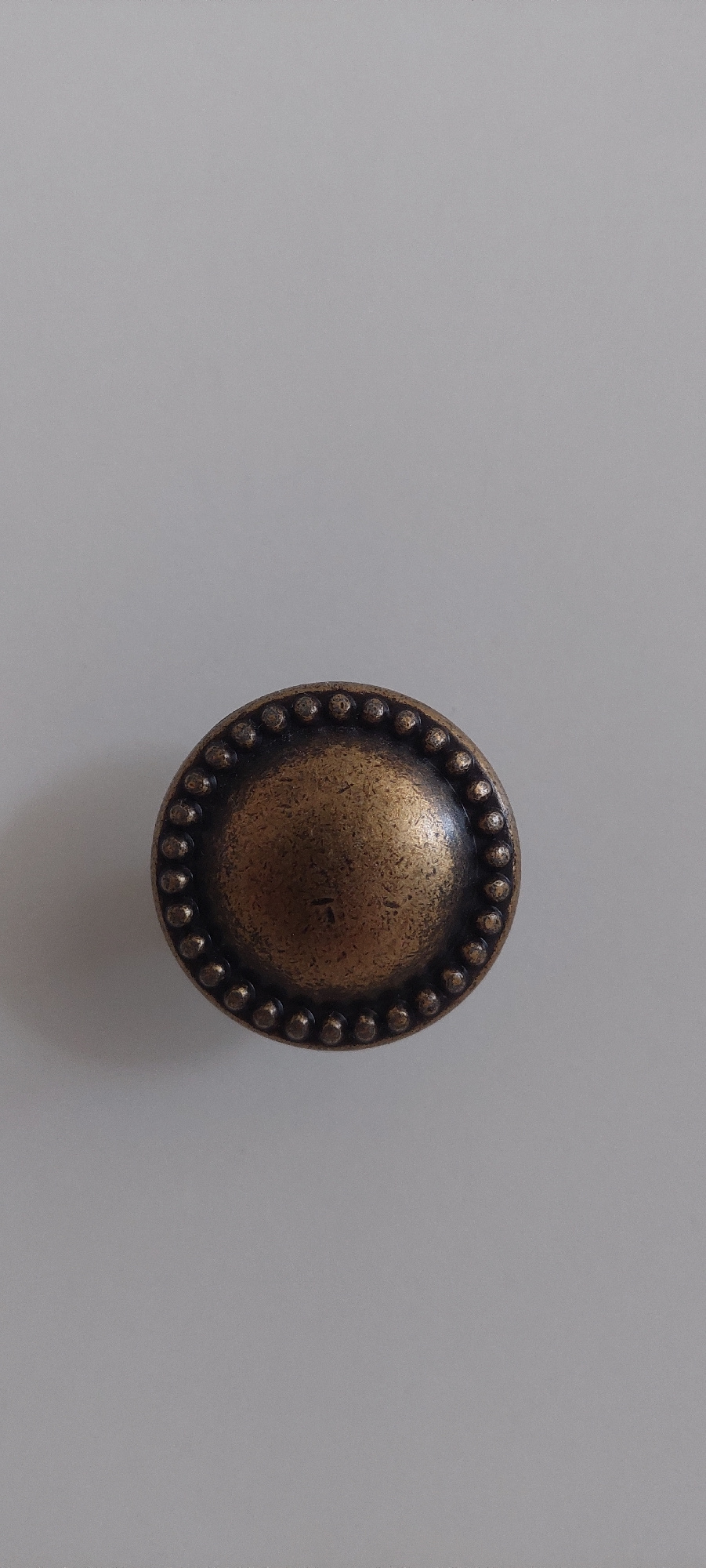 A circular, bronze-colored drawer pull with a beaded edge is centered on a plain, light gray background. The drawer pull has a slightly convex surface and is positioned horizontally within the frame. The background appears to be a smooth, matte surface, possibly a wall or a piece of furniture. There are no people or other objects visible in the image. The location of this object in the city of Ivanovo, Russia, cannot be determined from the image itself.
