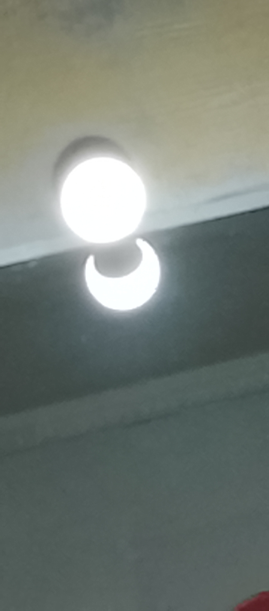 The image shows a bright, circular light source, likely a ceiling light fixture, positioned in the upper portion of the frame. Below and slightly to the right of the main light, there is a distinct crescent-shaped reflection or shadow, illuminated by the main light. The background appears to be a textured surface, possibly a ceiling, in a pale, yellowish-beige color, transitioning to a darker, grayish-blue surface below. This lower surface is likely a wall or another part of the room, and it creates a strong contrast with the illuminated elements above. The lighting is direct and intense, causing some overexposure on the main light source and a clear, sharp reflection.