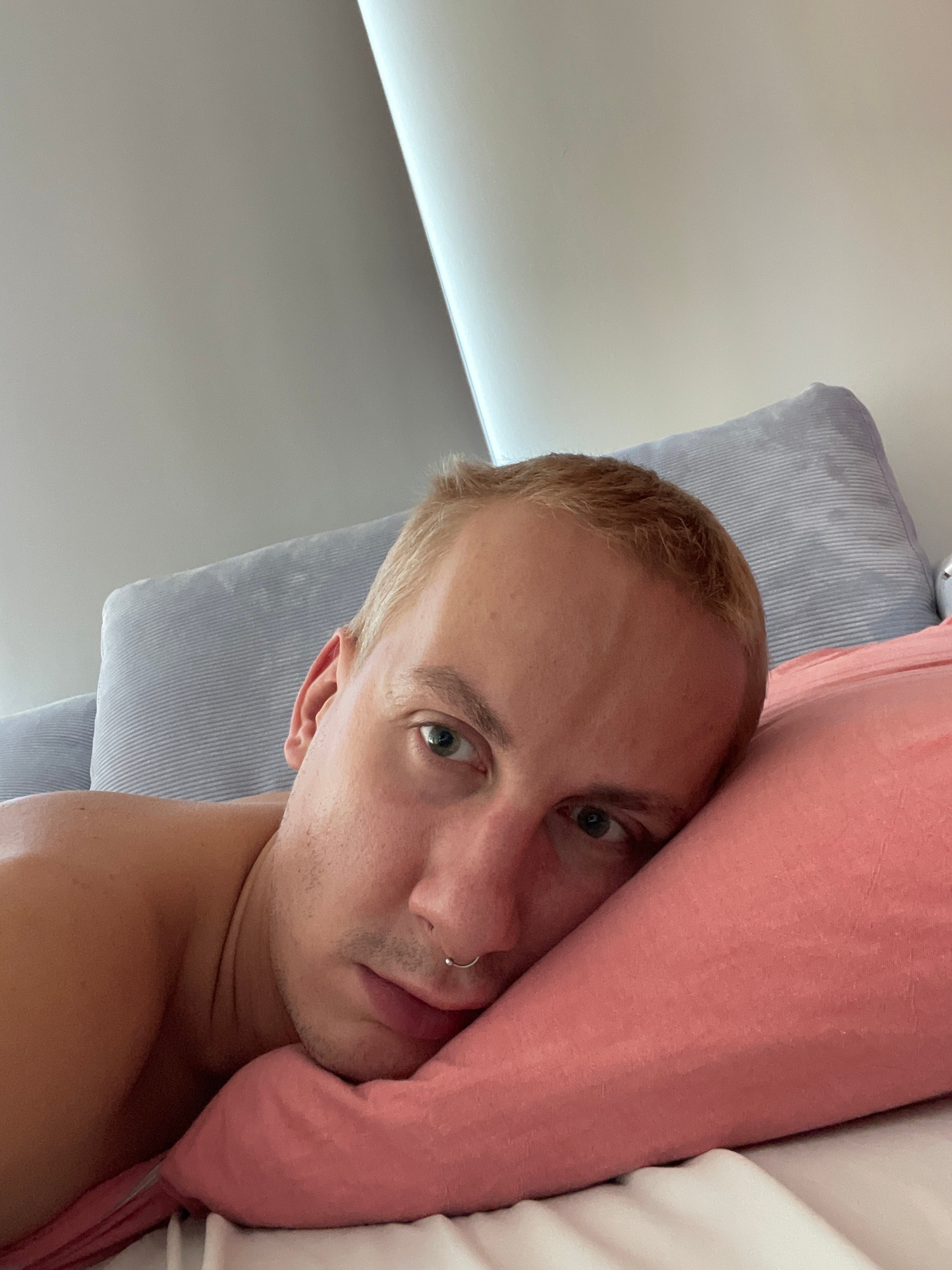 An eye-level, close-up, head-and-shoulders shot shows a light-skinned person with short, blonde hair and piercing blue eyes, looking directly at the camera. They are lying on their side, resting their head on a pink pillow, with their left shoulder and arm visible. A silver septum piercing is visible on their nose. Behind them, two blue-striped pillows are stacked. The background consists of a light grey wall and a white ceiling. The scene appears to be an indoor setting, possibly a bedroom. There is no indication of the location being in the city of Unknown, Russia.