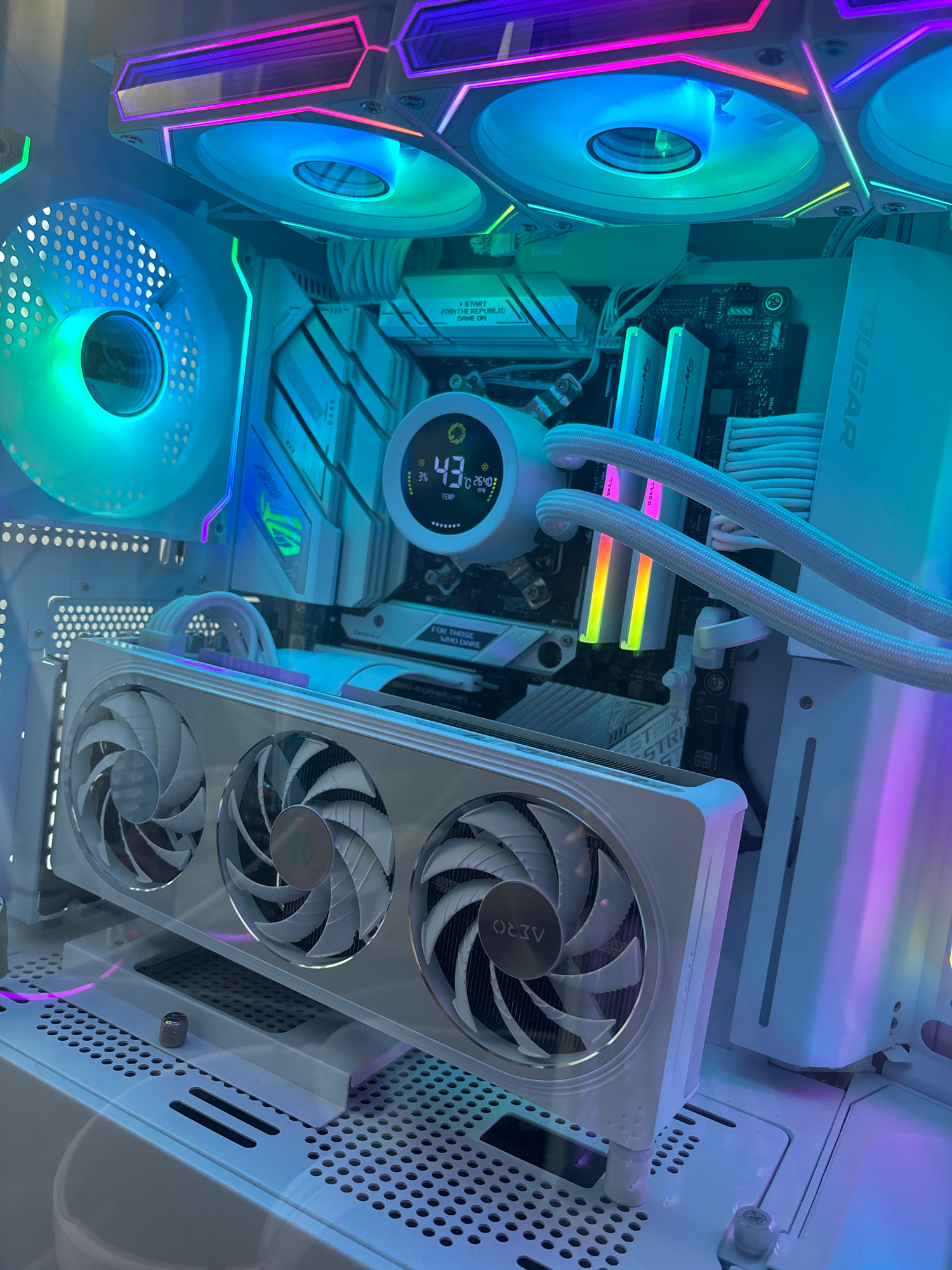 A white computer case is filled with internal components. The computer has several fans, including a large one on the left side and three fans on a white graphics card at the bottom. The components are lit by colorful RGB lighting, casting blue, purple, and green hues throughout the case. A small circular digital display shows "43°C" and "2640 RPM". The overall environment is the interior of a desktop computer, likely in a home or office setting, with no observable city or location context.