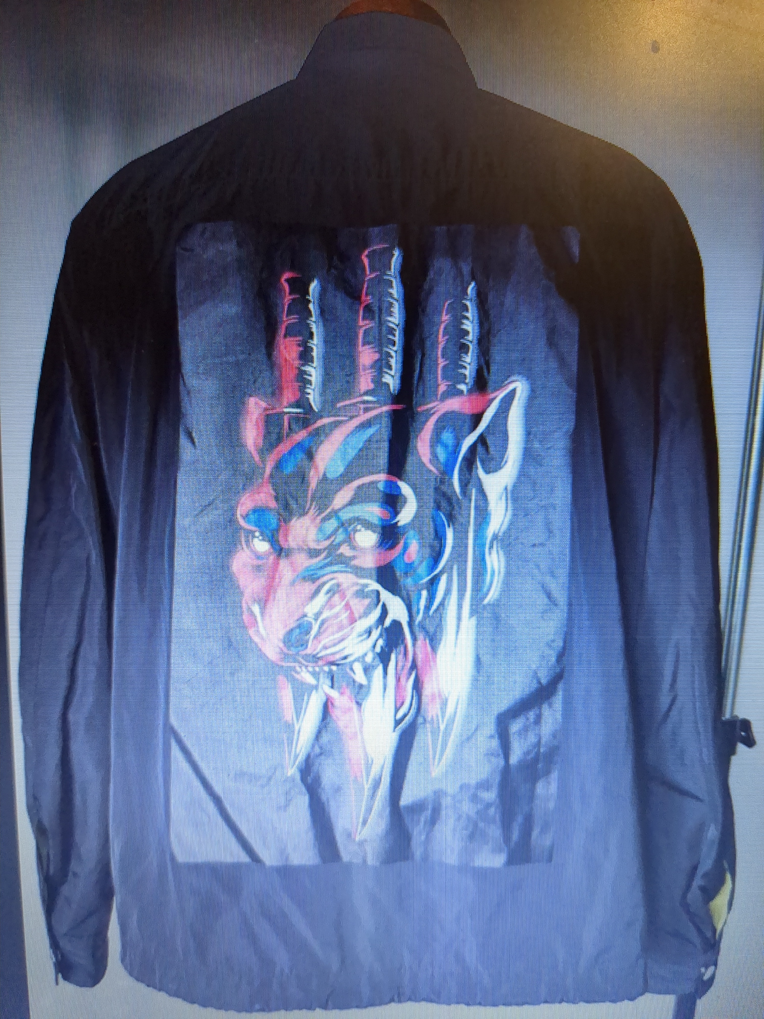 A black jacket is displayed. The back of the jacket features a large graphic print of what appears to be a stylized wolf's head with three swords piercing through it. The graphic uses vibrant colors, including red, blue, and white. The jacket is made of a lightweight, possibly nylon, material, and has long sleeves. There are no people present in the image. The background is a neutral, somewhat textured wall, and the jacket is likely displayed indoors, possibly on a hanger or mannequin, though the support structure is not fully visible. The image is taken from a slightly elevated, head-on perspective. There is no indication of the location being Minsk, Belarus.