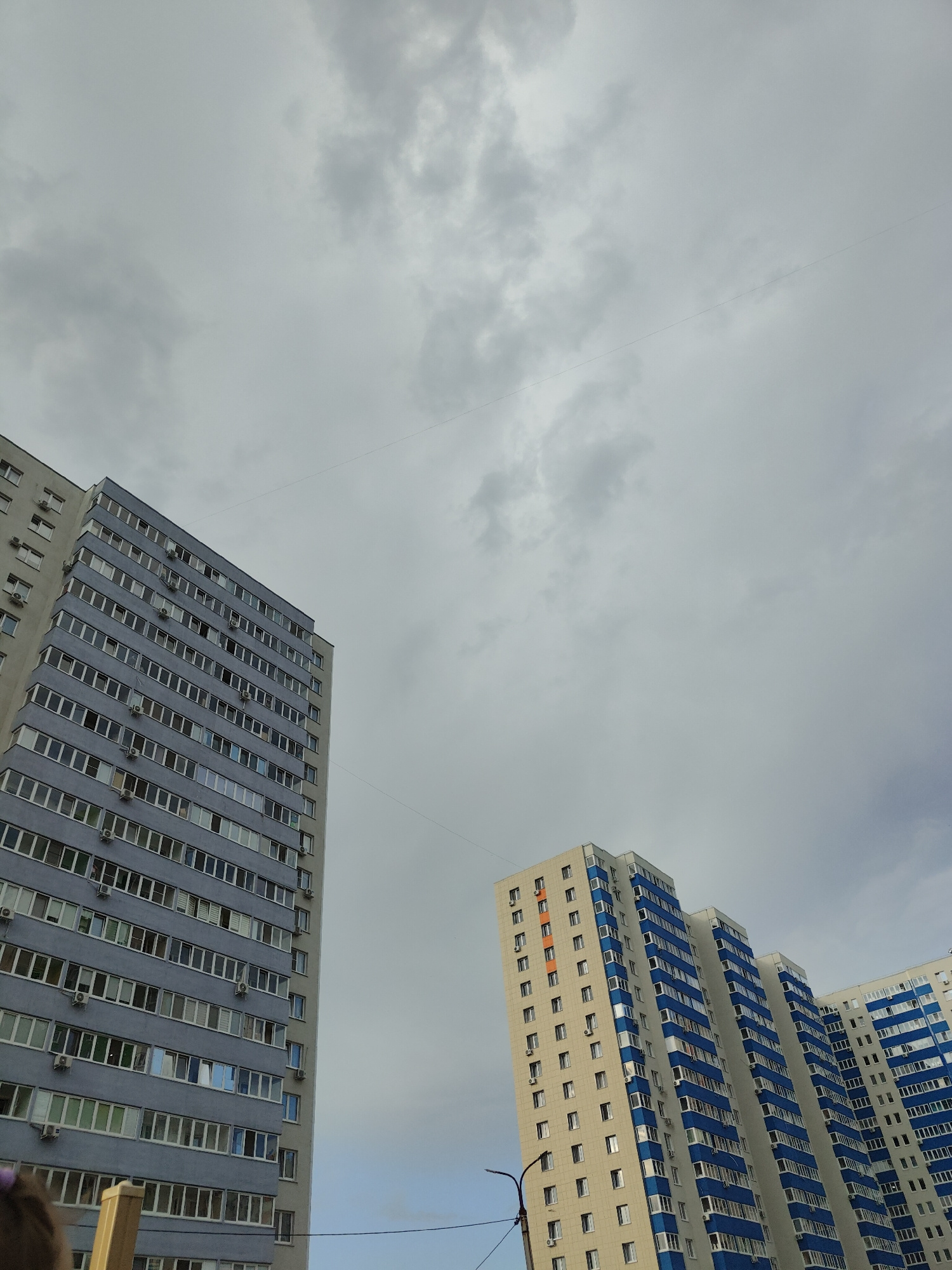 The image captures a view of several high-rise residential buildings against a cloudy sky. Two prominent buildings are in focus, viewed from a low angle looking upwards. The building on the left is tall and appears to be light gray with numerous windows, some of which are tinted blue. Air conditioning units are visible mounted outside many of the windows. The building on the right is a cluster of connected towers, primarily cream-colored with striking blue accents around the balconies and window frames. It also features many windows and air conditioning units. A thin, dark line, likely a wire, stretches diagonally across the sky. Another building, mostly obscured by the foreground structure on the right, is also visible. A street lamp is partially visible in the lower right portion of the image. The sky is overcast with varying shades of gray clouds, suggesting a cloudy or possibly rainy day. No people are clearly discernible, though a small, dark object, possibly part of a person's head or clothing, is visible in the bottom left corner. The overall scene suggests a residential urban environment in Ufa, Russia, likely during the daytime. There is no visible text in the image.