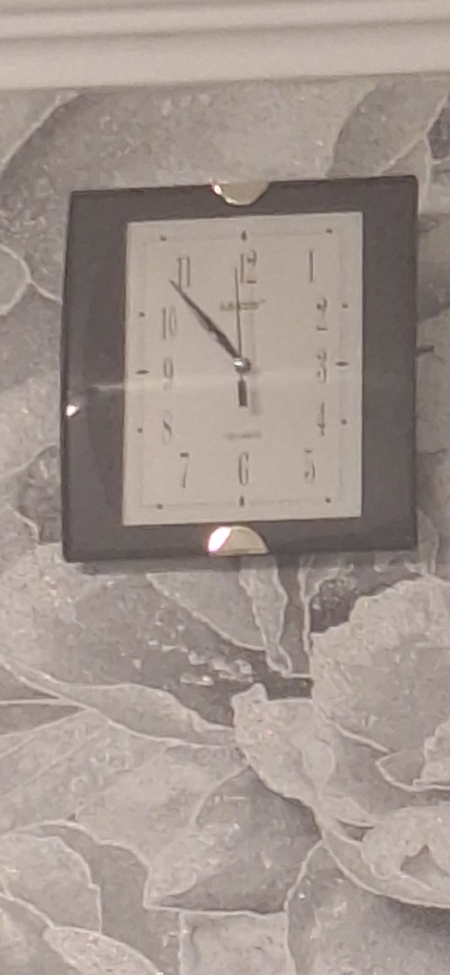 A square wall clock is mounted on a textured wall. The clock face is white with black numerals and hands. The hour hand is pointing to approximately 10, and the minute hand is pointing to approximately 3. The clock has a dark gray frame. A bright light is reflecting off the bottom edge of the clock frame. The wall has a light gray, textured pattern resembling floral or abstract shapes. There is a white molding at the top of the wall. The image is taken from a slightly low angle. The location of this image is not provided.