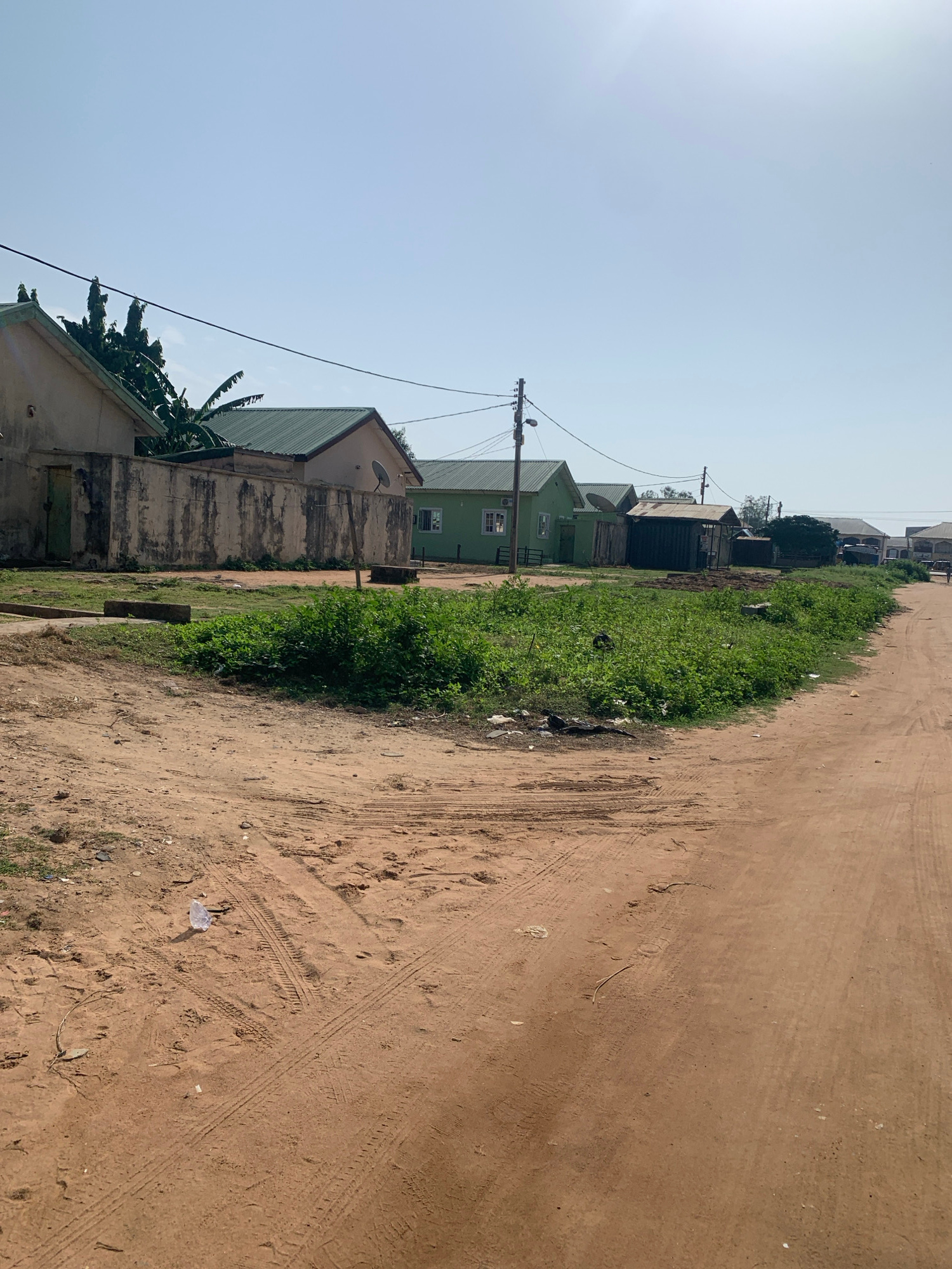 The image depicts a dusty, unpaved road winding through a residential area. On the left side of the road, a row of houses with green roofs is visible, separated by a weathered concrete wall. Lush green vegetation grows in front of the houses. The road itself is marked with tire tracks and scattered litter, including a plastic bag. In the distance, more houses and a few trees can be seen under a clear blue sky. The scene suggests a warm, possibly arid climate.
