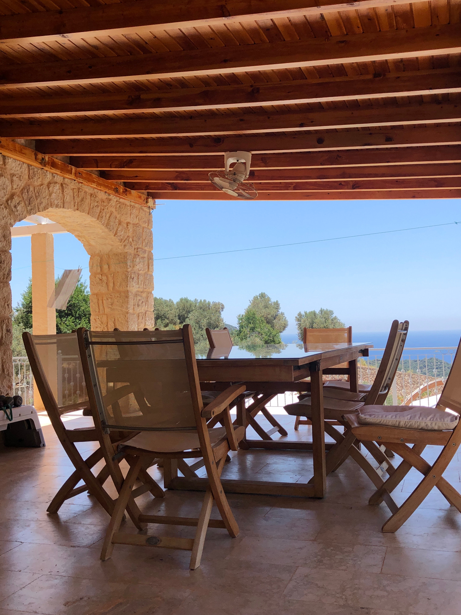 This is an outdoor shot taken from a covered patio overlooking the sea. The patio has a wooden slatted ceiling and stone pillars. A wooden table with a glass top is set with several wooden chairs, some of which have light-colored cushions. In the background, lush green trees and the blue sea are visible under a clear sky. The location is the city of Kaş, Turkey.