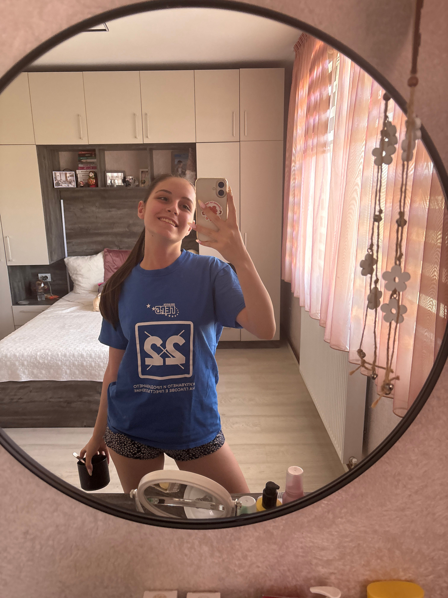 A young woman with long brown hair pulled back into a ponytail takes a selfie in a large oval mirror. She is wearing a royal blue t-shirt with a white and blue logo on the front and a black and white patterned skirt. The logo on her t-shirt appears to be in Cyrillic script, reading "SS" within a square border. The text below the logo also appears to be in Cyrillic. She is smiling and holding her phone in her right hand, with her left arm slightly raised.
The mirror reflects a bedroom scene. In the background, a bed with white bedding and a pink pillow is visible. To the left of the bed, there are tall white cabinets with silver handles. To the right of the bed, there is a nightstand with a lamp and some other items. The walls are a light beige color with a textured finish. Above the cabinets, there is a window with sheer pink curtains. A string of decorative flower-shaped ornaments hangs from the ceiling in front of the curtains. The overall lighting suggests it is daytime, possibly indoors with artificial lighting. The scene appears to be a personal space, likely a bedroom in a home. The woman's expression is happy and relaxed.