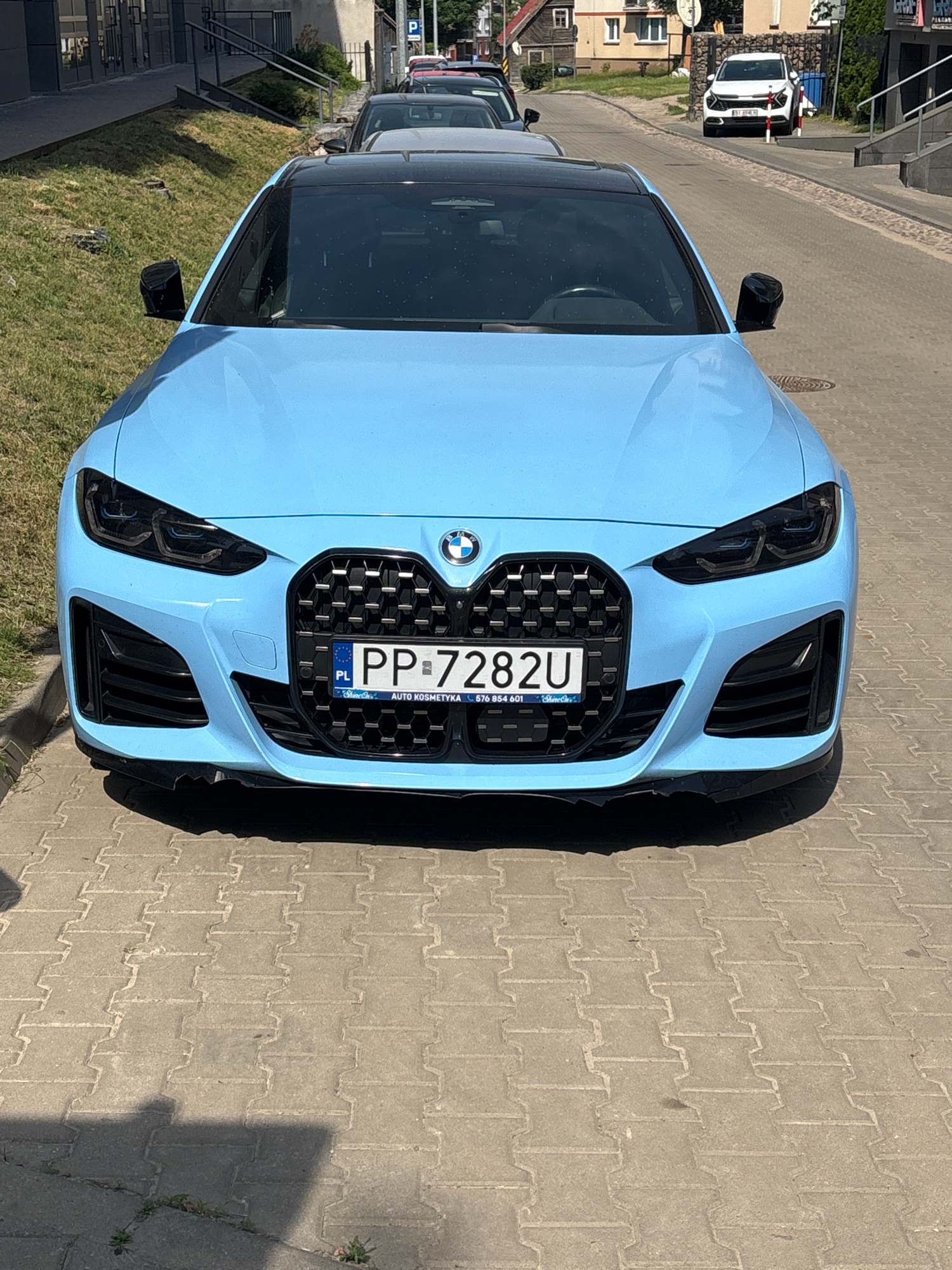A light blue BMW is parked on a paved street, viewed from the front. The car has a black grille with a honeycomb pattern and black headlights. A Polish license plate reading "PP 7282U" is mounted on the front of the car. Behind the BMW, other cars are visible, parked in a row. To the left of the BMW, there is a grassy area and a metal railing. In the background, buildings and trees can be seen. The scene appears to be a street in Białystok, Poland. The lighting suggests it is daytime, likely a sunny day given the shadows cast by the car. The pavement is made of interlocking concrete blocks. There is no visible activity or people in the image.