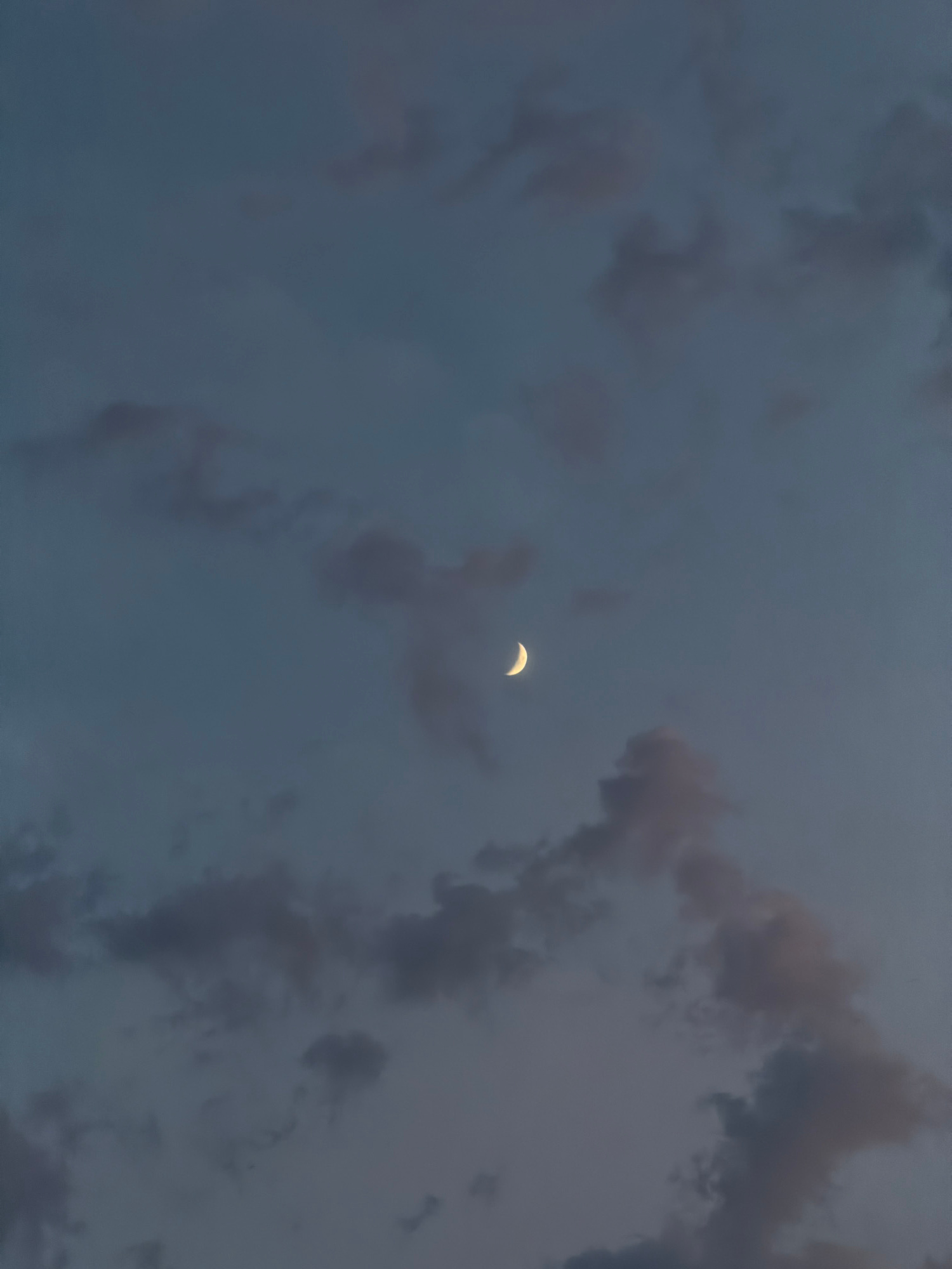 A waxing crescent moon is visible in a dusky, cloudy sky. The clouds are scattered and appear to be dark grey with hints of purple and orange, suggesting either dawn or twilight. The moon is a bright, thin sliver of light against the darker backdrop. The scene is devoid of any human presence, structures, or specific landmarks that would indicate a particular location within the city of Tuapse, Russia. The focus is solely on the celestial event and the atmospheric conditions.