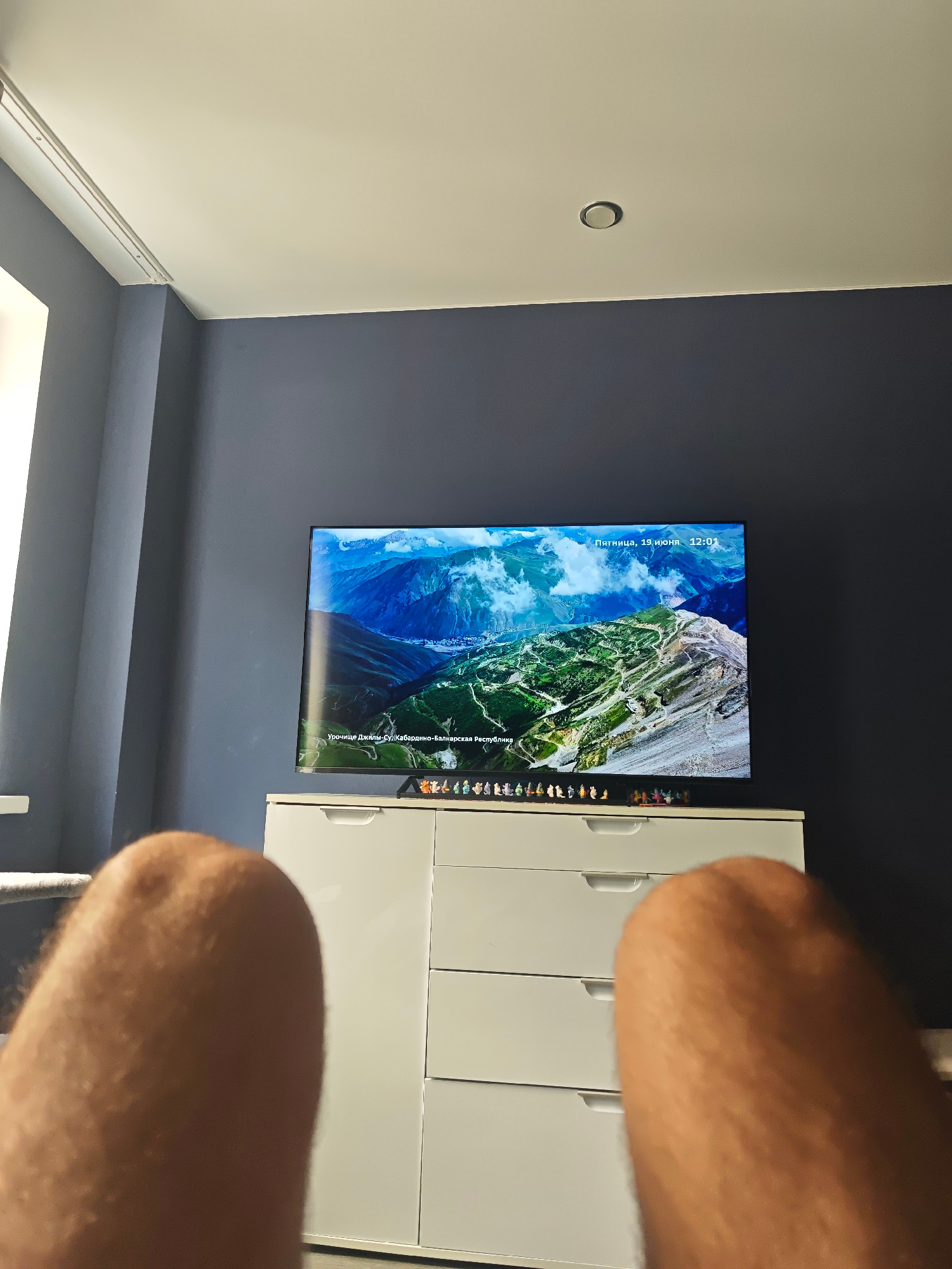 An interior shot captures a television displaying a mountainous landscape with a date and time overlay indicating "Friday, June 19, 12:01". The TV is placed on top of a white dresser with several drawers. The wall behind the dresser is painted a dark blue. In the foreground, blurred legs are visible, suggesting a person is sitting or lying down facing the television. The scene is likely in a residential setting, possibly a living room. The image does not provide any information that can be used to determine a location in the city of Razdolnoye, Russia.