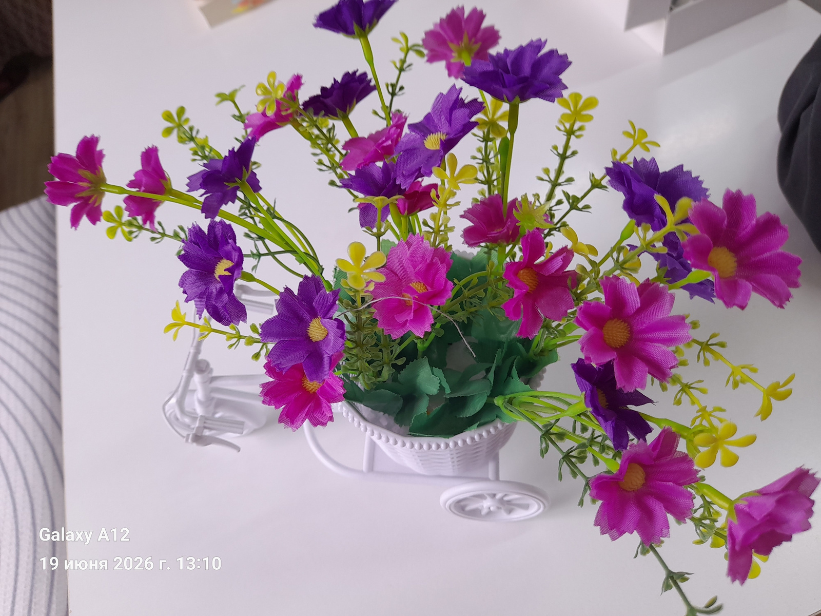 The image displays a decorative arrangement of artificial flowers in a white, bicycle-shaped planter. The flowers are a mix of vibrant pink and purple with small yellow accents. The planter itself is designed to resemble a miniature bicycle with wheels and handlebars, all in white. The arrangement is placed on a white surface, likely a table or shelf, with a blurred background that includes a textured fabric and a hint of a white ceiling. There is no discernible activity taking place, and the lighting suggests an indoor setting.
Visible text in the lower left corner of the image reads "Galaxy A12" and "19 июня 2026 г. 13:10". This indicates the photograph was taken with a Samsung Galaxy A12 phone on June 19, 2026, at 1:10 PM. The location context provided is Kara-Köl City, Kyrgyzstan.