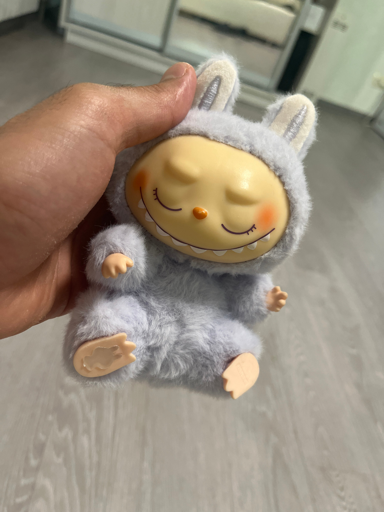 An overhead, medium shot captures a hand gently holding a plush, pale blue bunny-eared toy. The toy has a round, yellow face with closed eyes, rosy cheeks, and a wide, toothy smile with visible white teeth. Its body is covered in soft, fuzzy blue material, with small tan hands and feet. The background is blurred, showing a light-colored, wood-grain floor. The scene appears to be indoors, in a domestic setting, potentially a home in Novokuybyshevsk, Russia, given the requested location context, although no specific landmarks or urban features are visible to confirm this.