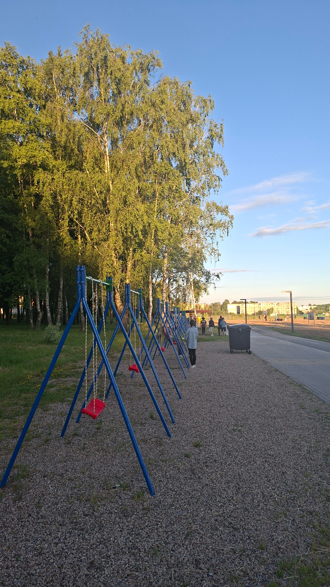 The image depicts a playground with multiple blue A-frame swing sets, each featuring a red swing seat. The swings are positioned on a gravel surface adjacent to a paved walkway. In the background, a line of trees, predominantly birch trees with their distinctive white bark, forms a dense foliage backdrop. Beyond the trees, a clear blue sky is visible with a few wispy clouds. On the paved walkway, several people are walking away from the camera, some in small groups. A large, dark grey trash bin is situated near the walkway. In the far distance, buildings and streetlights suggest an urban or suburban setting. The scene appears to be set in the late afternoon or early evening, with soft, natural lighting illuminating the environment. Based on the user's prompt, this scene is located in Smalyavichy, Belarus.