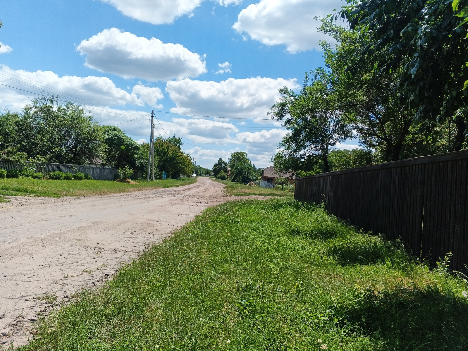 A slightly elevated, wide-angle, eye-level shot captures a rural dirt road in an unspecified location on a clear day. The road, appearing somewhat uneven and cracked, curves gently towards the distance, flanked by lush green grass on the right and a wooden fence on the left. The left side of the road also features a wooden fence and scattered trees. Overhead, a vibrant blue sky is dotted with large, fluffy white clouds. Power lines stretch across the scene, supported by utility poles. A few houses and trees are visible in the background. The overall impression is one of a serene, undeveloped rural or suburban environment.
