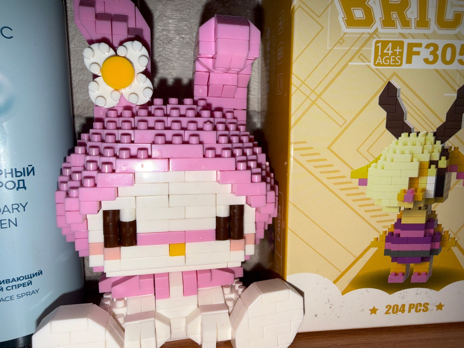 The image shows a close-up of several objects on a flat surface, likely a table or shelf. On the left is a light blue cylindrical bottle with Russian text that appears to be a cosmetic product. To the right of the bottle is a pink brick toy figure resembling a character with large ears and a flower accessory on its head. Next to this toy is a yellow box with graphics and text, including the number "F305" and "14+ AGES". On the box, there is another small brick toy figure of a yellow and purple creature with horns and a striped torso. The bottom of the box has text that reads "★ 204 PCS ★". The overall lighting is dim and appears to be artificial. The scene is composed of these three items and their immediate surroundings. There is no visible indication of the city of Rayevskiy, Russia, or any specific location details.
