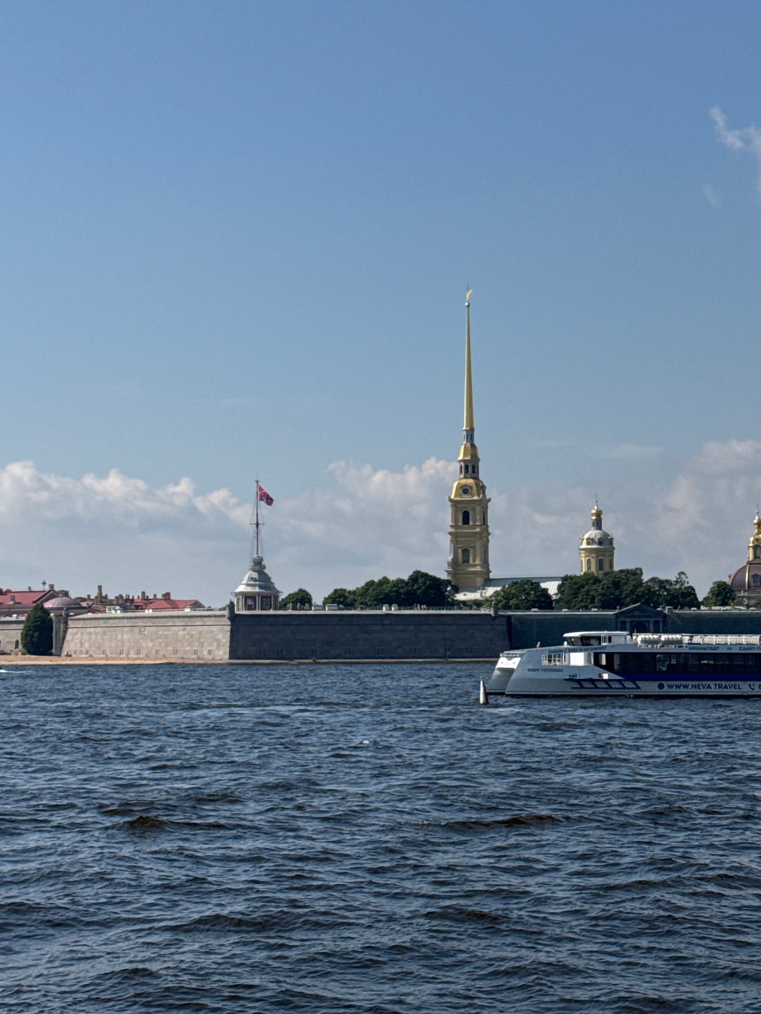 This image shows a scenic view of Saint Petersburg, Russia. In the foreground, a dark blue body of water with moderate waves fills the bottom of the frame. A white tour boat with blue accents is traveling on the water, its side displaying the text "WWW.NEVA.TRAVEL".
In the middle ground, the Peter and Paul Fortress, a prominent landmark in Saint Petersburg, is visible along the waterfront. The fortress features a long, stone wall with a series of windows. A tall, slender spire topped with a golden angel stands out as the most distinctive element of the fortress's architecture, characteristic of the Peter and Paul Cathedral within the fortress. Adjacent to the spire are other smaller domed buildings, also typical of Russian Orthodox churches. A red flag flies from a flagpole near the fortress walls. Green trees are planted behind the fortress walls, providing a contrast to the stone structures.
The background consists of a clear blue sky with scattered white clouds, suggesting a pleasant day. The overall scene captures a view of the Neva River with the historic Peter and Paul Fortress on its banks.