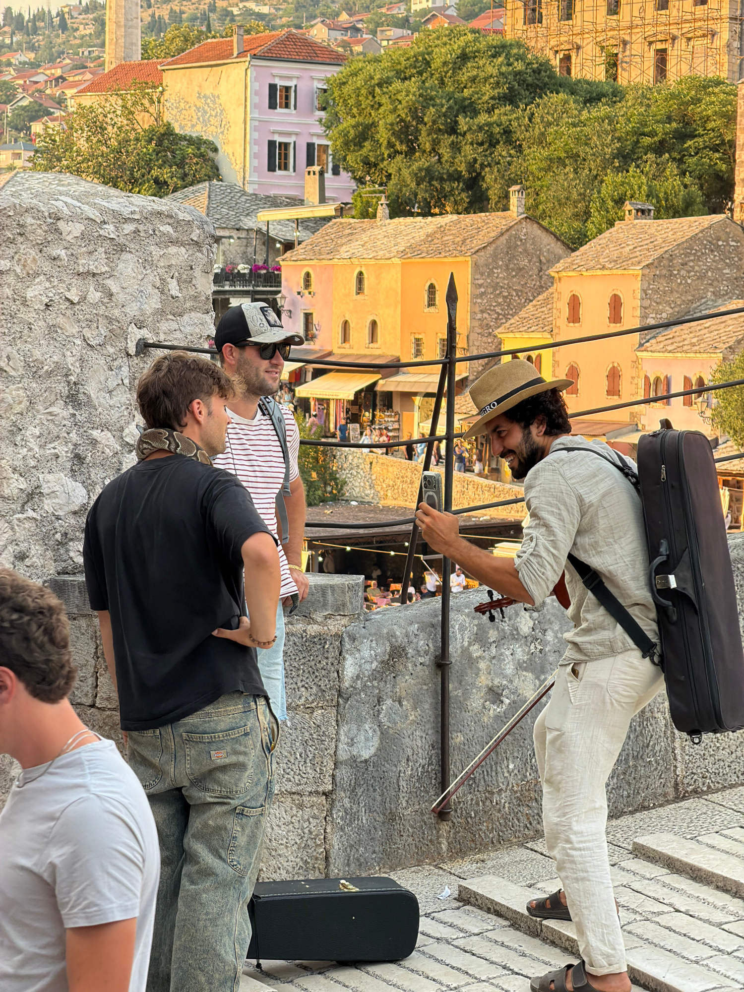 A snake handler with a python wrapped around his neck stands with two other men on a stone wall overlooking a city with terracotta roofs. One man wears a black t-shirt and distressed jeans. The second man wears a striped t-shirt, a baseball cap, and sunglasses. The third man, a street performer, wears a straw hat and a linen shirt, holding a violin and bow, and taking a selfie with his smartphone. He has a violin case strapped to his back. Buildings with colorful facades and pitched roofs are visible in the background. The scene appears to be on the Old Bridge in Mostar, Bosnia and Herzegovina, given the architecture and the vista.
