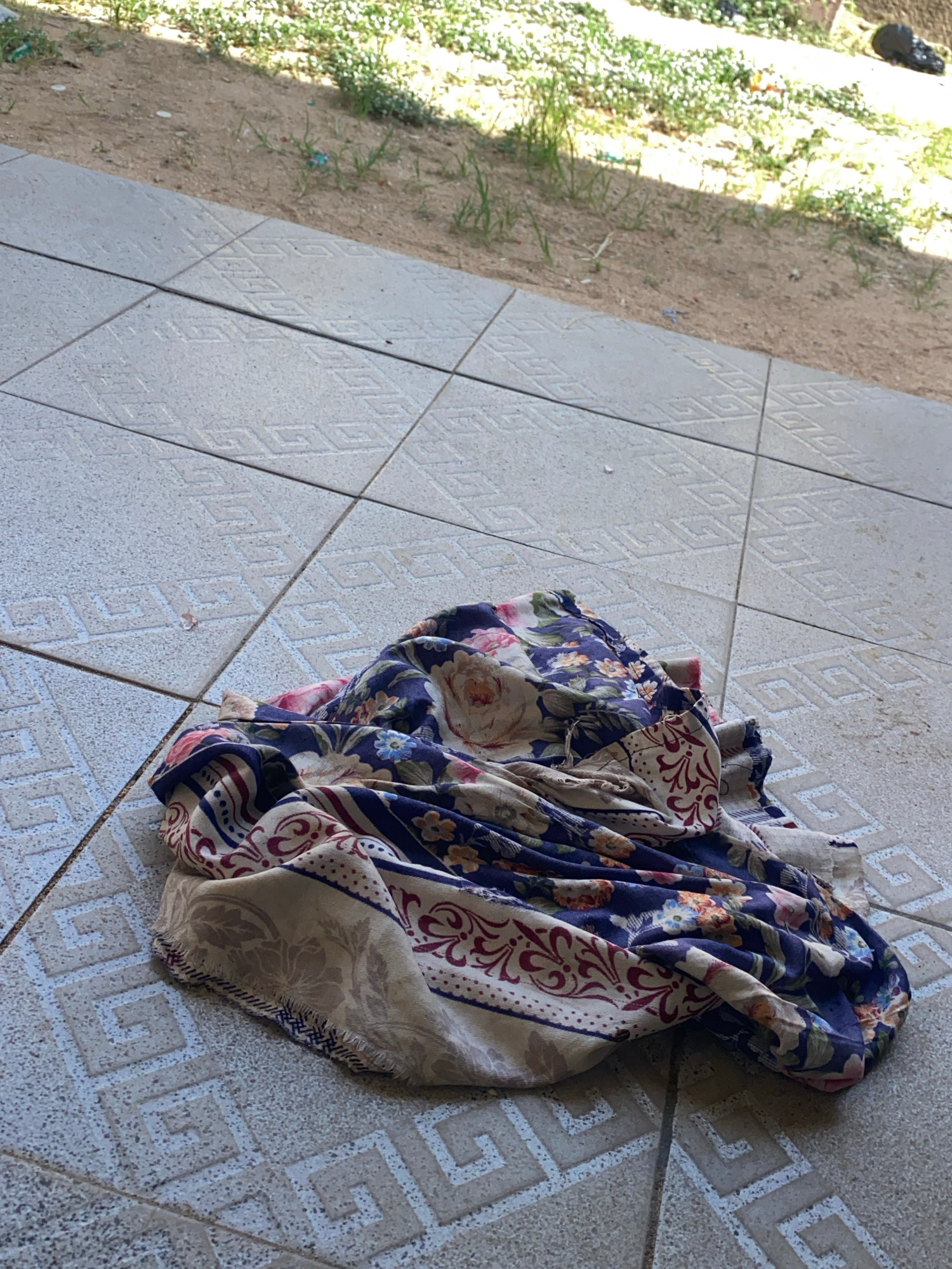 A floral-patterned fabric is draped on a tiled surface. The fabric features a dark blue background with pink, white, and orange flowers, interspersed with green leaves. A border with a cream and burgundy pattern runs along one edge. The tiled surface is made of light-colored square tiles with an embossed geometric pattern. Behind the tiles, there is a patch of dry, sandy ground with sparse green vegetation. The scene appears to be outdoors, possibly in a courtyard or patio area.