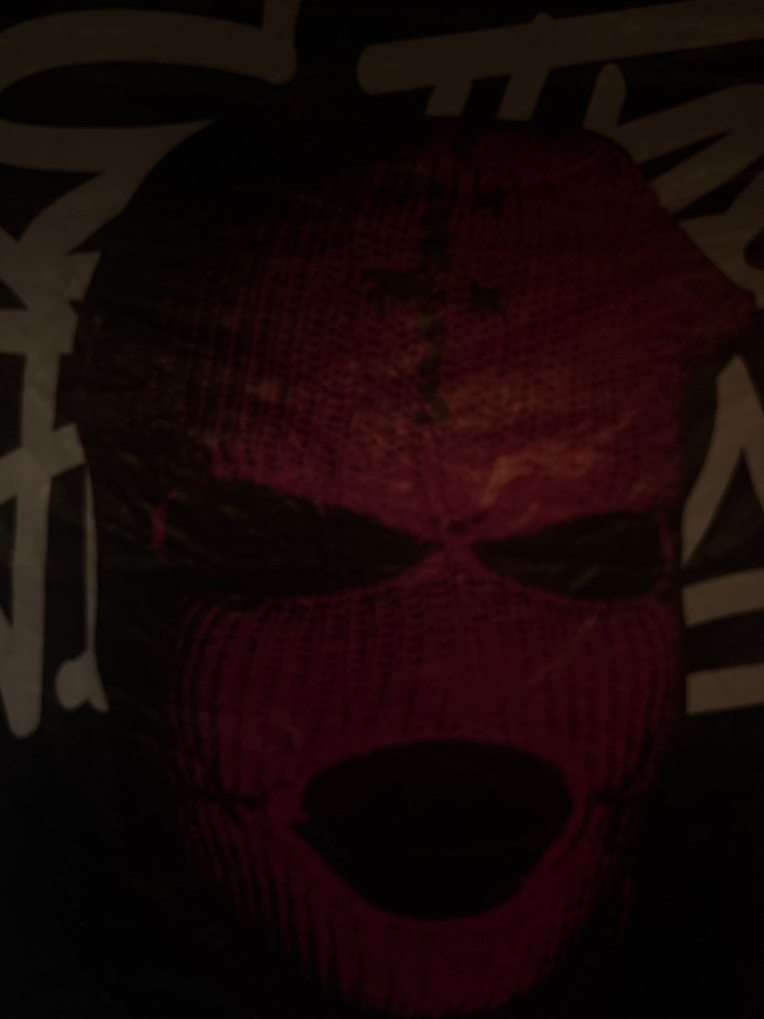 The image is a close-up, blurry shot of a red knitted ski mask with black eye holes and a gaping mouth opening. The mask appears to be part of a garment or design, possibly a t-shirt, with abstract white lettering visible in the background. The lighting is dim, casting shadows and obscuring fine details. The overall impression is dark and slightly ominous. The location is Unknown, Unknown.
