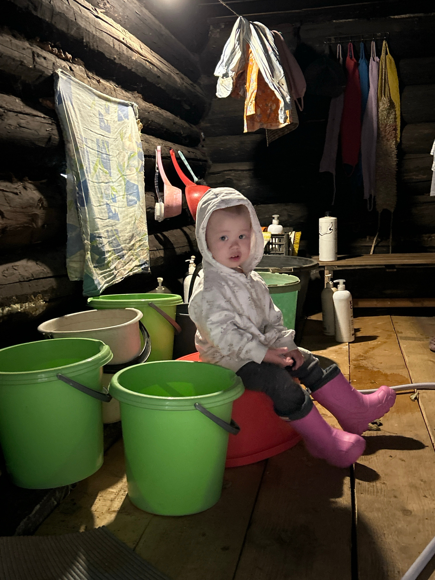 In a rustic, dimly lit room constructed of dark logs, a young child wearing a white hooded jacket and pink rubber boots sits on a red plastic basin. The child is surrounded by several green plastic buckets, one of which is larger and partially visible in the foreground. To the left, a patterned towel hangs from the wall. In the background, various items are hanging from a rack on the wall, including clothing and what appear to be bath accessories. On a wooden shelf behind the child, there are bottles, including a pump dispenser bottle and a roll of toilet paper. The floor is made of wooden planks. The scene suggests a bathroom or utility area within a log cabin. The location is identified as Азинский, Russia.