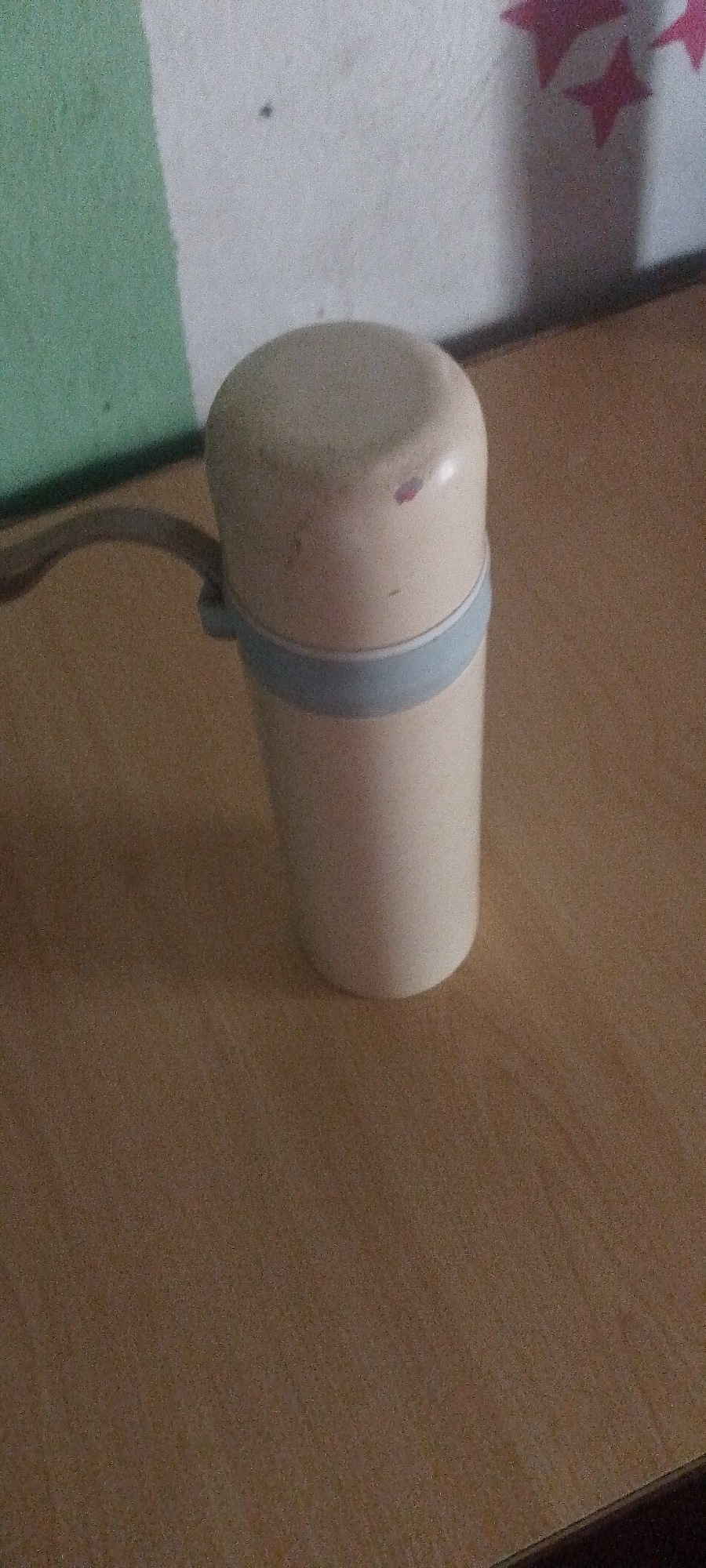 A light beige, cylindrical object with a rounded top and a light blue band around its middle sits on a wooden surface. The object appears to be a dispenser or a small container. A dark, coiled cord extends from the left side of the object and curves behind it. The background consists of a wall that is partially green and partially white, with some pink star-shaped markings on the white section. The lighting is dim, casting shadows and highlighting the textured surface of the wooden table. The image is grainy and lacks sharp focus, suggesting it was taken with a low-resolution camera or in poor lighting conditions.