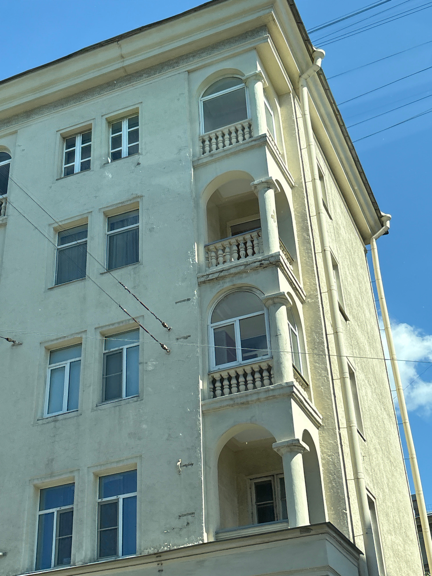 This image shows the side of a light-colored, multi-story building with a rounded balcony on the second floor, located in Saint Petersburg, Russia. The building has a weathered facade with visible stains and cracks. There are multiple windows on each floor, some with white frames and panes, others with darker frames and obscured views inside. Decorative elements such as arched window frames and balustrades on the balcony are visible. Overhead, power lines are strung across the blue sky, with some clouds present. The building appears to be part of an urban environment, possibly residential or office space, given its architecture and the presence of utility infrastructure.