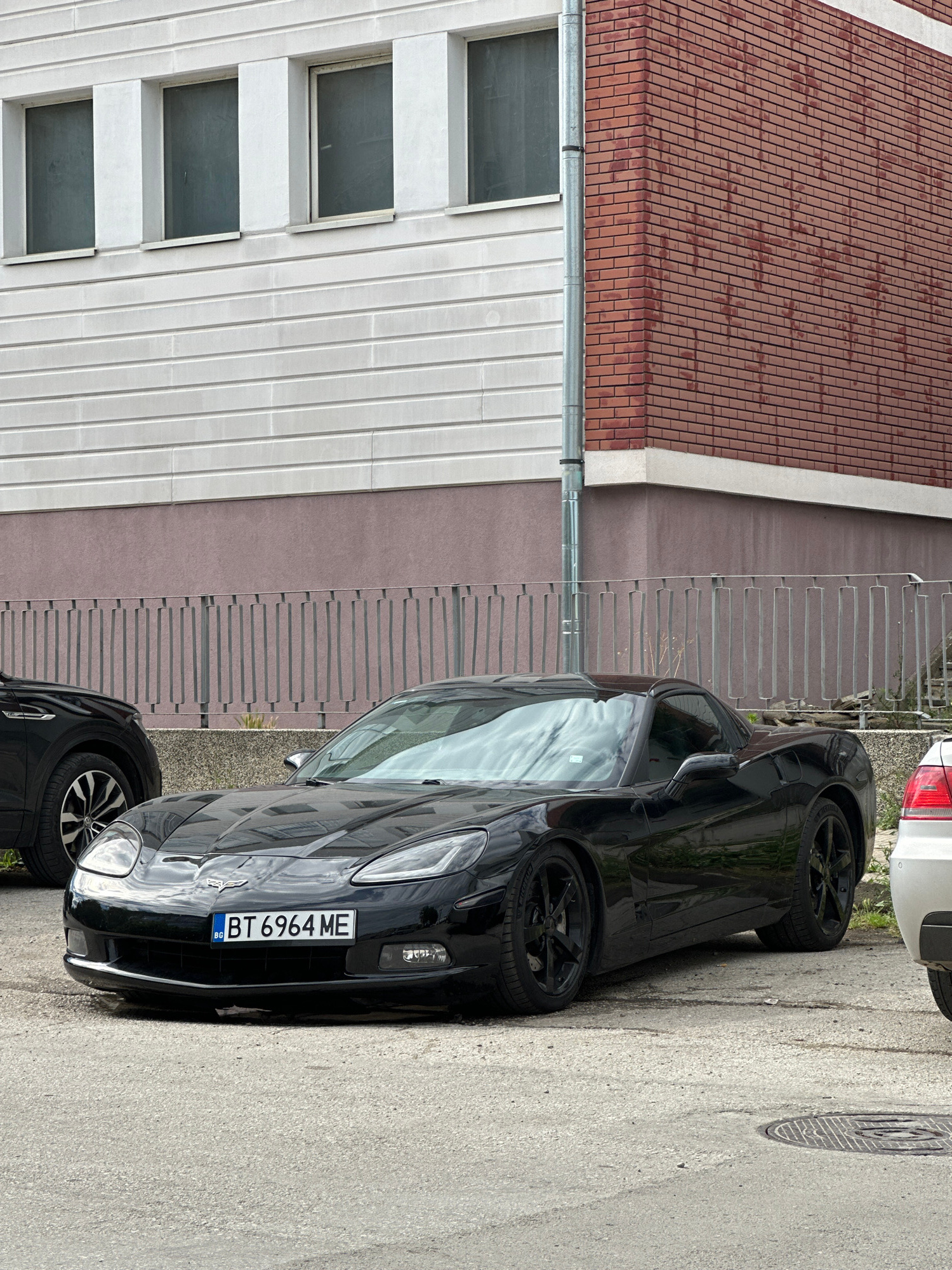 A black Chevrolet Corvette is parked on a paved surface in front of a building in Veliko Tarnovo, Bulgaria. The building has a facade of white horizontal siding and a section of red brick. A metal fence runs along the front of the building. The Corvette has a license plate that reads "BT 6964 ME". Another car is partially visible on the left, and a white car is visible on the right. The sky is overcast and reflected in the car's windshield.