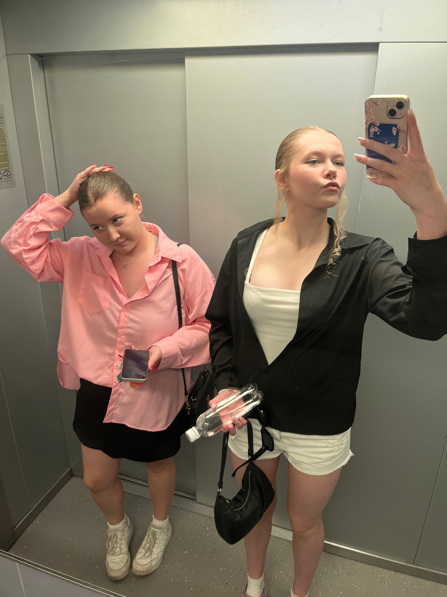 Two young women are taking a mirror selfie in an elevator. The woman on the left is wearing a pink button-down shirt and a black skirt. She has her hand on her head and is holding a smartphone in her other hand. The woman on the right is wearing a white tank top, a black open shirt, and white shorts. She is holding a bottle of water and a smartphone in her hands, and is making a pouty face towards the camera. The elevator appears to be in a modern building. The location is not identifiable as Perm, Russia.
