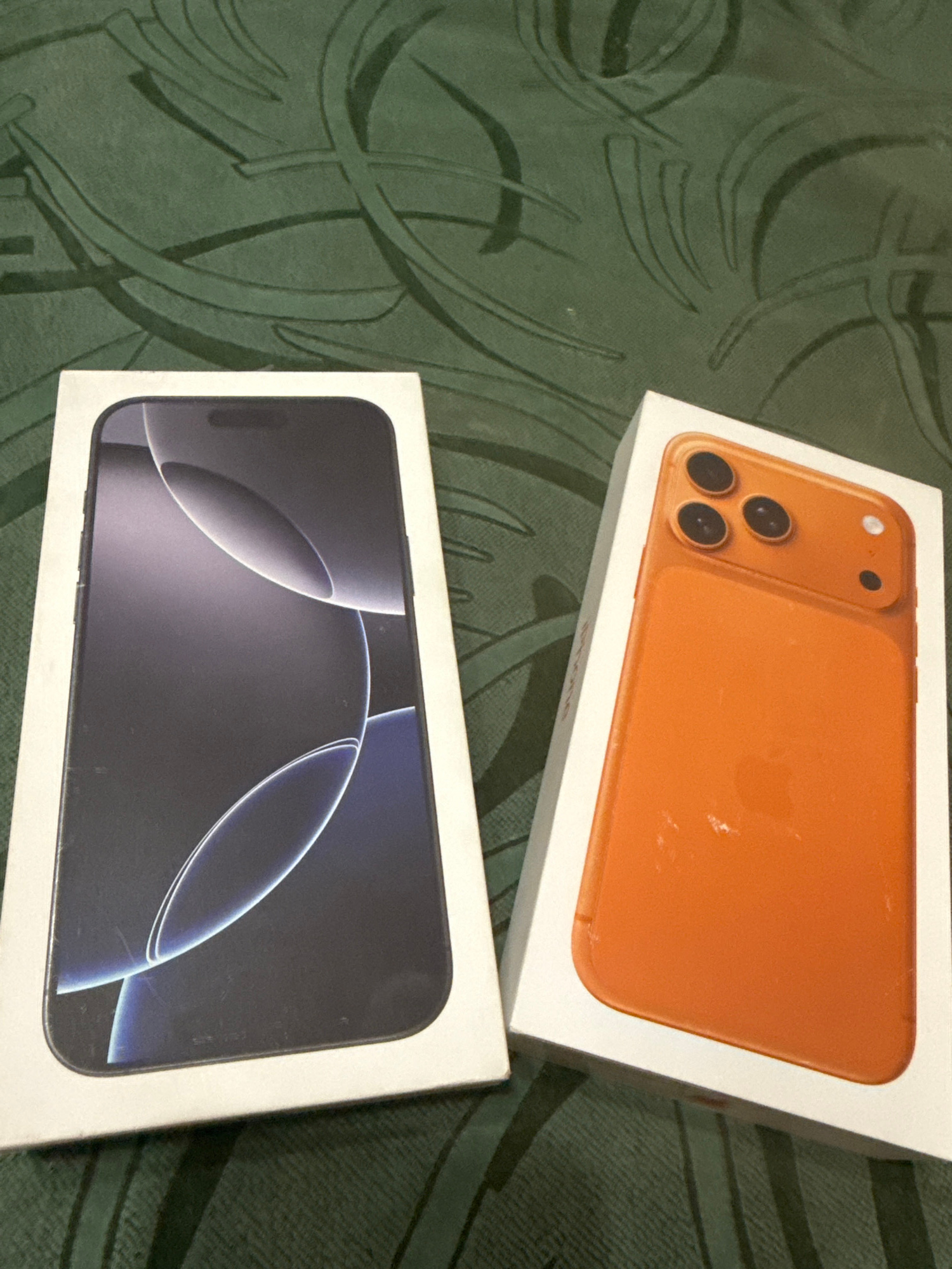 Two iPhone boxes are placed on a green, patterned surface. The box on the left displays a dark-colored iPhone with a prominent camera cutout. The box on the right shows an orange-colored iPhone, also with a visible camera module. The background is a dark green fabric with a repeating leaf-like pattern. The scene appears to be indoors, on a flat surface, likely a table or a bed. There are no people or animals present in the image.