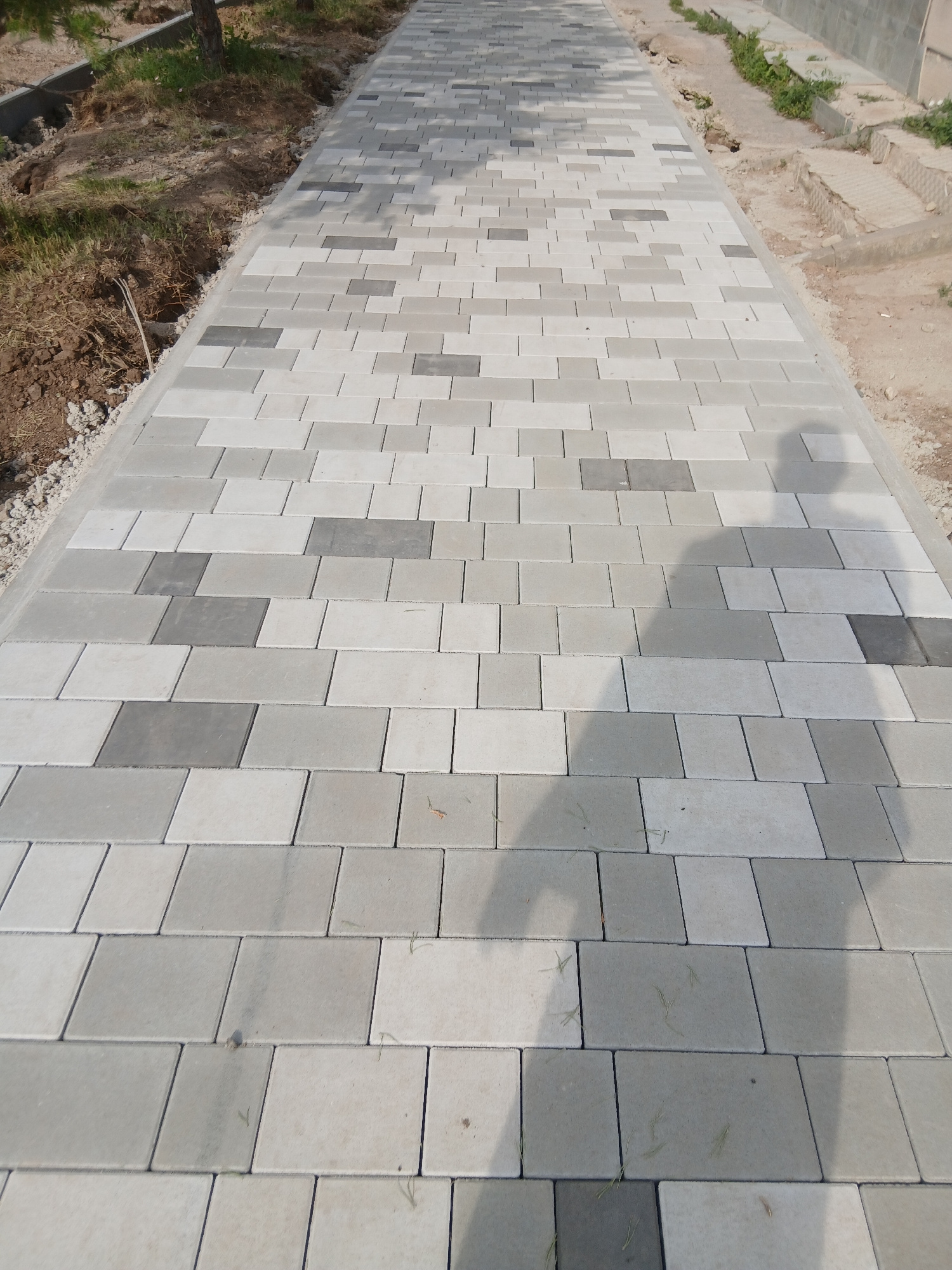 The image depicts a newly paved walkway with rectangular interlocking concrete pavers in varying shades of grey and white. The walkway extends into the distance and is bordered on the left by a patch of dry soil, grass, and a tree trunk. On the right, the walkway is adjacent to a dirt area with some construction debris and a portion of a concrete structure. A shadow of a person is visible on the walkway, suggesting it is daytime. The scene is likely a public space or park in Astana, Kazakhstan, given the recent construction activity indicated by the fresh paving and surrounding dirt.