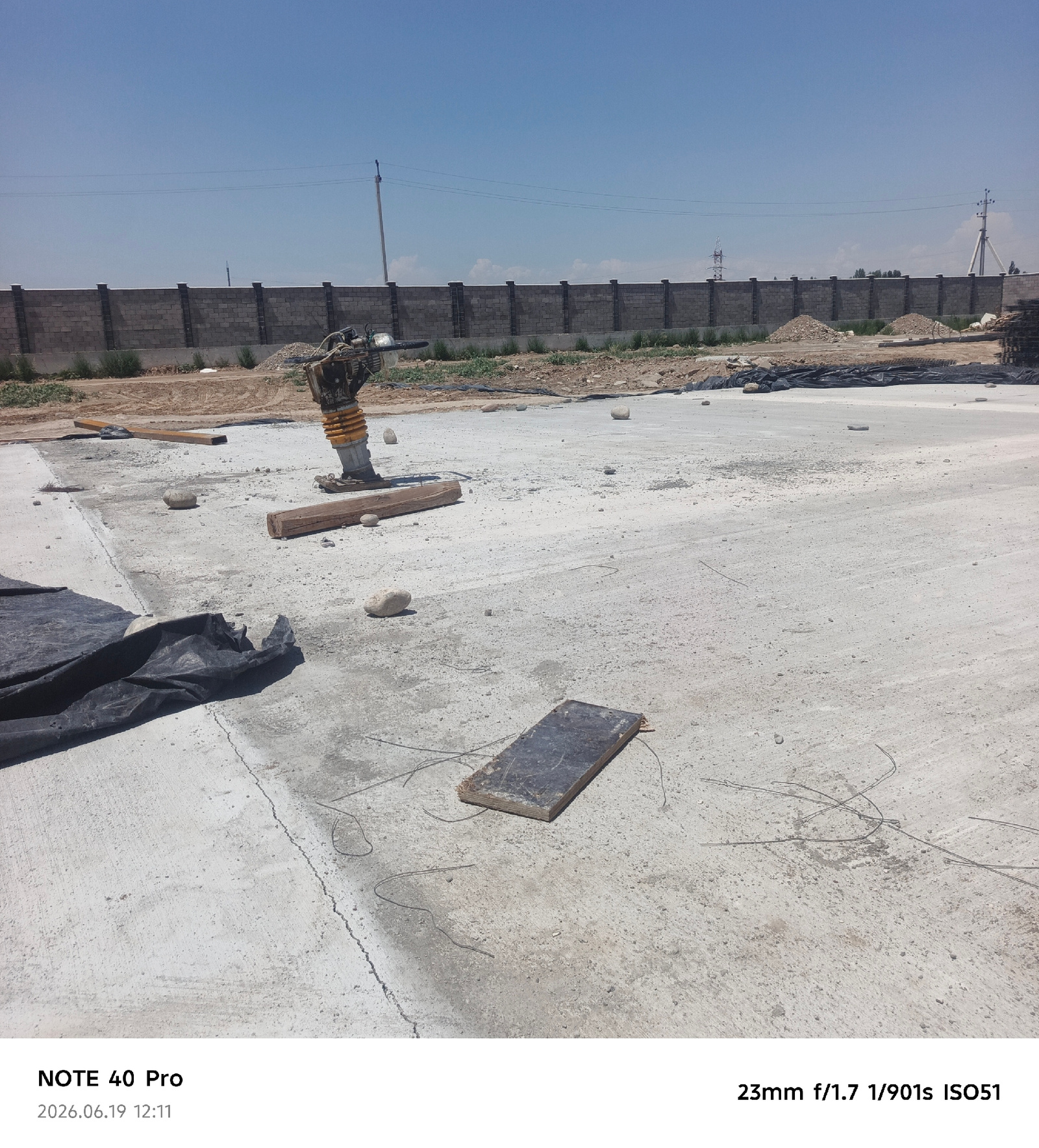 The image captures a construction site under a clear blue sky. In the foreground, a concrete surface is visible, with scattered stones and a black tarp. A piece of dark wood, possibly a plank, lies on the concrete. To the left, a vibratory compactor, yellow and black, stands on the surface, with wooden beams nearby. In the middle ground, more concrete and dirt are present, along with piles of construction debris. A grey brick wall with dark posts forms a boundary in the background, with sparse vegetation in front of it. Beyond the wall, power lines stretch across the sky, and a distant pylon can be seen. The image is dated 2026.06.19 12:11 and includes technical details: 23mm f/1.7 1/901s ISO51. The overall scene suggests ongoing construction work in an open area.