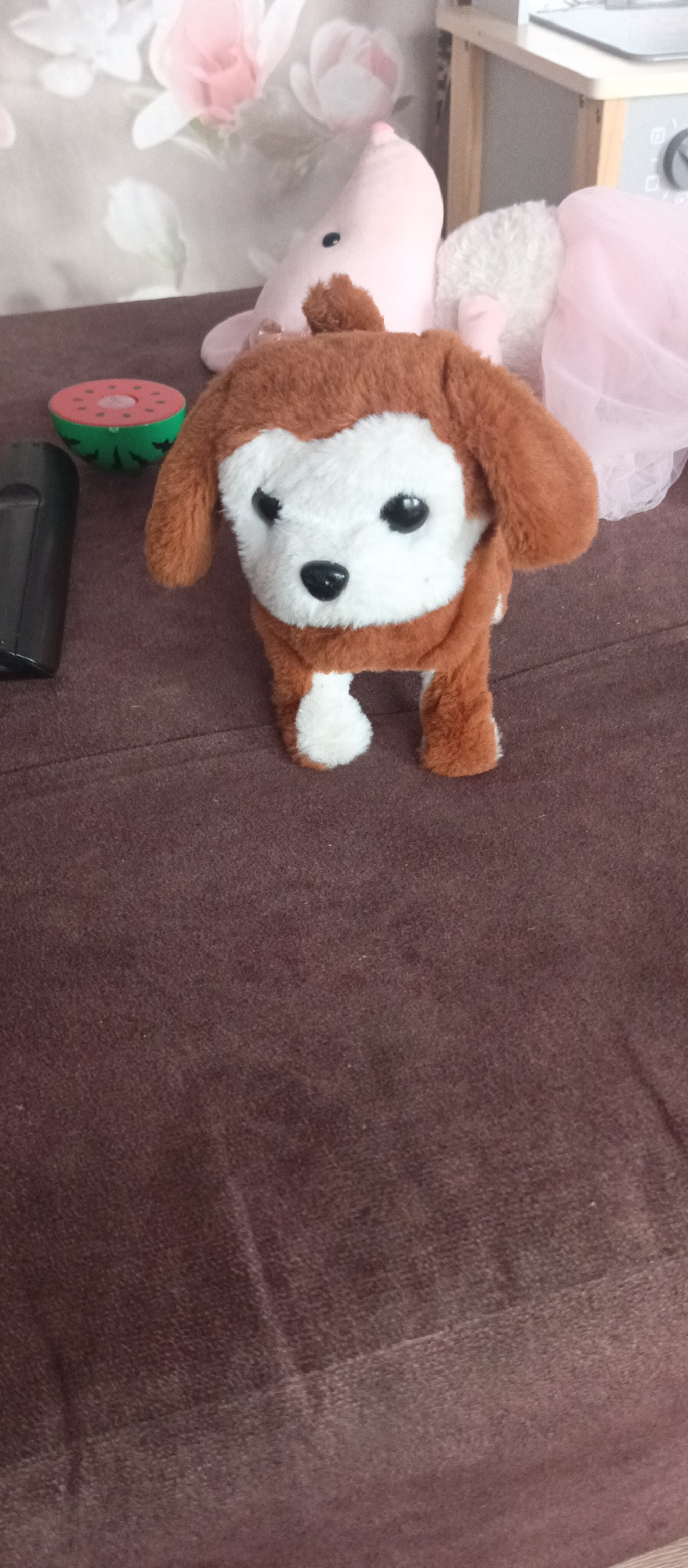 In this close-up image, a stuffed toy dog with brown fur and a white face is the central subject. It stands on a textured, brown surface, likely a couch or rug. The toy has black button eyes and a black nose. To the left of the dog, a slice of a toy watermelon with a green rind and red interior is visible. In the background, there are blurred floral patterns on what appears to be wallpaper, with hints of pink and white flowers. Behind the dog and to the right, parts of other soft toys are visible, including a pink pig-like toy with a black eye, and a fluffy white toy. A sheer pink fabric is also draped in the background. The overall impression is one of a child's play area or a collection of stuffed animals. No people are visible, and the activity is static, focusing on the arrangement of the toys. There is no visible text. The lighting suggests an indoor setting during the daytime.