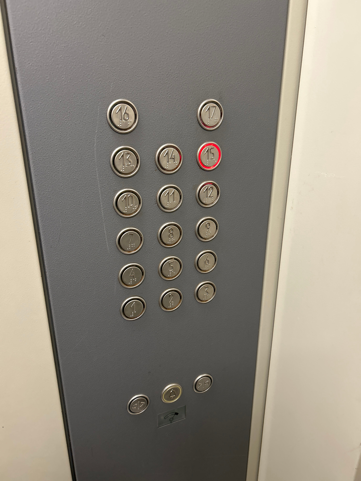 The image shows a close-up view of an elevator control panel. The panel is dark gray and features a grid of circular buttons, each with a number and Braille text. The buttons are arranged in columns and rows, with numbers from 1 to 17 visible. The button for floor 15 is illuminated in red. There are also two additional buttons at the bottom of the panel, one with an icon of an open door and another with a bell icon. The panel is mounted on a lighter-colored wall. The image is taken from a slightly elevated perspective, looking down at the panel.