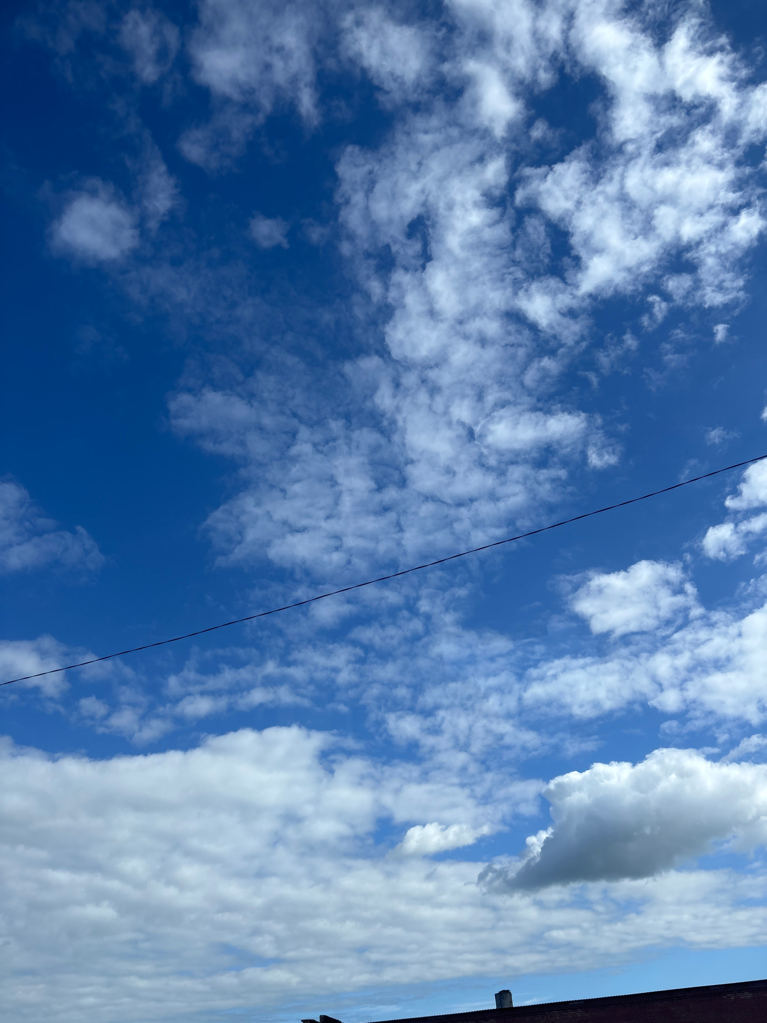 The image captures a bright blue sky filled with scattered white clouds. A thin, dark power line stretches diagonally across the upper half of the frame. In the lower portion of the image, the dark brown roof of a building is visible, with what appears to be a small, grey chimney stack. The scene is set in Kapyĺ, Belarus.