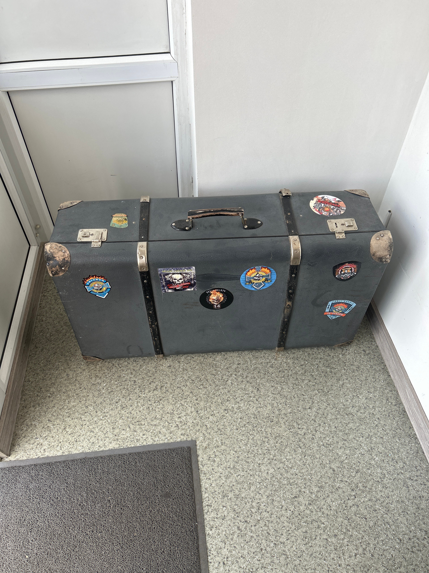 This media file shows a stack of three dark gray, rectangular suitcases placed on a speckled gray carpet. The suitcases have silver metal corners and latches. The top suitcase is adorned with several circular stickers, including one with an orange and yellow sunburst design. The middle suitcase also has stickers, one of which appears to be a skull. The bottom suitcase has a handle and a sticker with a blue and yellow shield. The background consists of a white wall and a light gray trim at the top. To the left of the suitcases is a white cabinet or door. The scene appears to be indoors, likely in a room with minimal natural light. There are no people or animals visible. The overall impression is one of storage or perhaps a travel-related display. The sticker text "BILA TSERKVA" is visible on one of the stickers, indicating the location as Bila Tserkva, Ukraine.