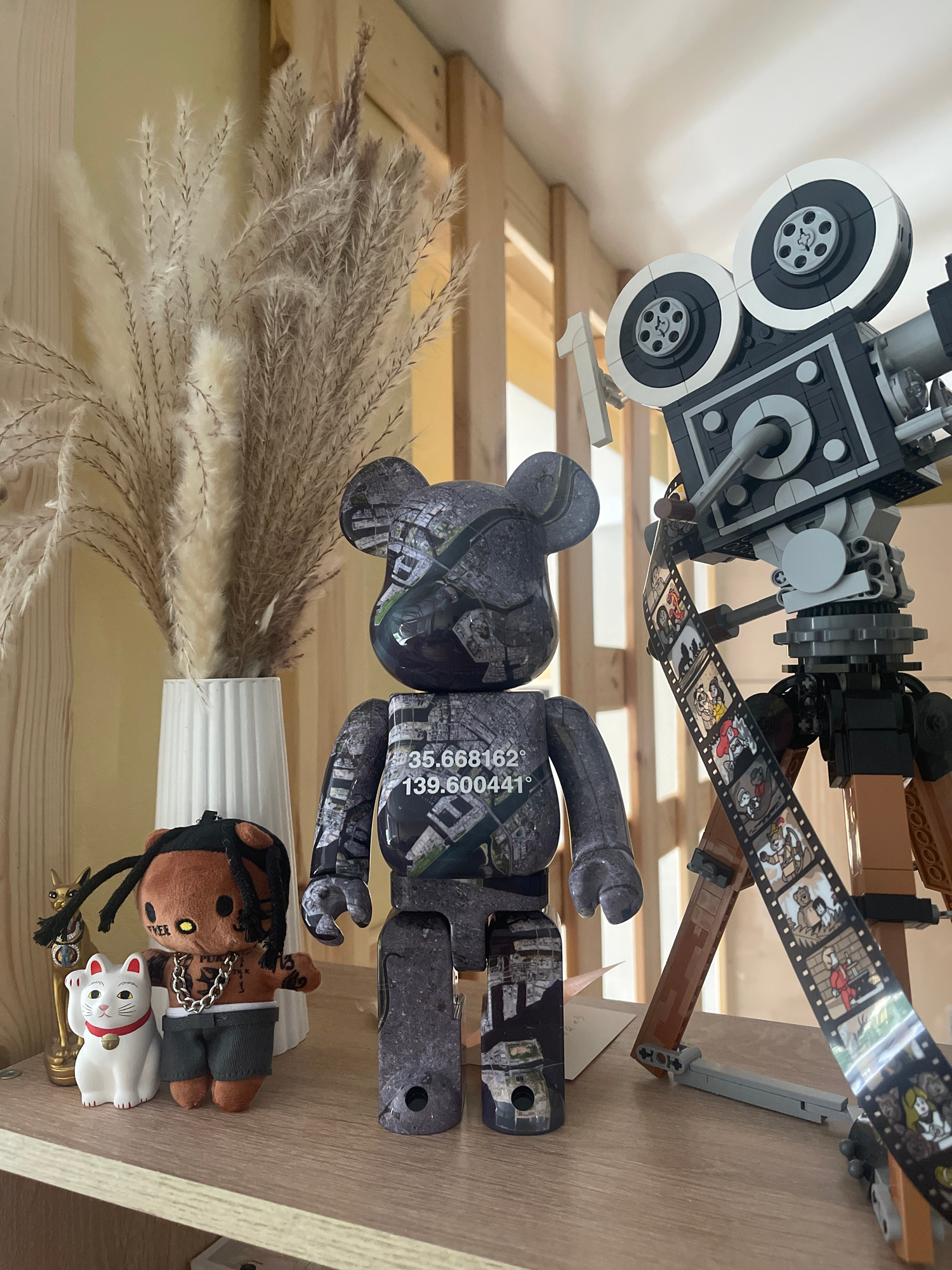 In an indoor setting, likely a shelf or display area, a collection of objects is arranged. The primary subjects are two large, dark gray bear-shaped figures, possibly figurines or collectibles, adorned with a city map pattern. One figure has coordinates printed on its arm: "35.668162" and "139.600441°". To the left of the bear figures is a bundle of dried, feathery reeds in a white vase. In front of the reeds sits a small plush toy resembling a person with dark hair, wearing gray shorts and a silver chain. Next to the plush toy is a small white ceramic cat with a red collar, a maneki-neko.
Behind these items, a black and gray Lego construction resembling an old-fashioned movie camera with film reels is prominently displayed. The film reel displays a sequence of cartoon-like images. The entire scene is set against a backdrop of wooden shelves, suggesting a home or retail display. The lighting appears to be artificial, casting soft shadows. No specific time of day or weather conditions can be determined.