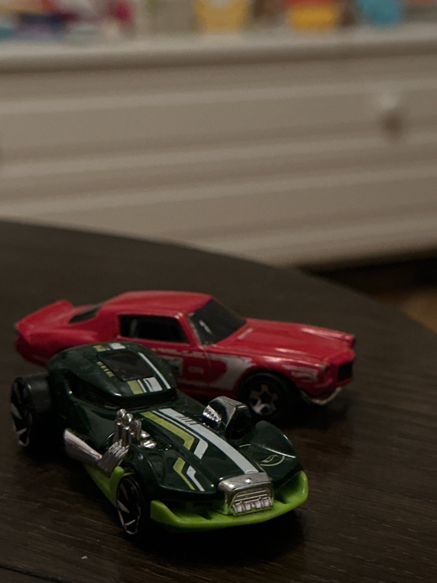 Two toy cars are on a wooden surface. The car in the foreground is dark green with lime green accents and white racing stripes. It has exposed silver engine parts and large black wheels with silver rims. The car in the background is red with white racing stripes and silver rims. The background is out of focus and appears to be a piece of furniture with various objects on top. There is no indication of the city of Sofia, Bulgaria.