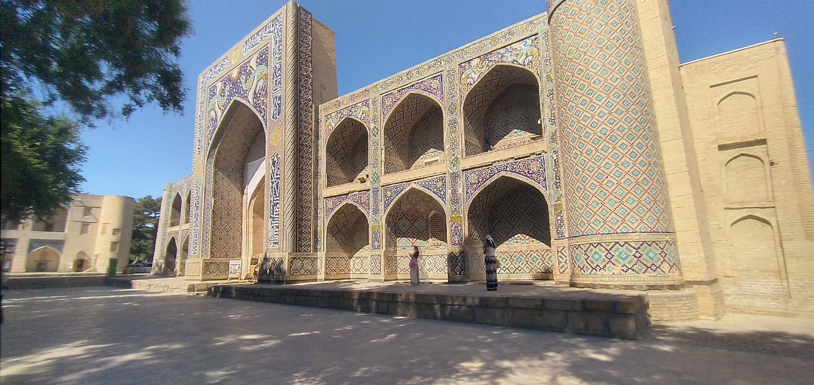 The image shows a large, ornately decorated building with multiple arched entrances and windows, characteristic of Islamic architecture, with intricate tilework featuring geometric patterns and stylized motifs. Two individuals are visible standing on a raised platform in front of the building. The building is made of light-colored brick, with significant portions covered in colorful tiles in shades of blue, turquoise, yellow, and white. The architecture suggests it is a madrasah or a mosque. The scene is set in a courtyard paved with gray stones, with shadows cast by an unseen tree on the left. In the background, another building with a rounded tower and arched windows is partially visible. This building is located in the city of Bukhara, Uzbekistan, likely part of the historic Lyab-i Hauz complex, which includes several madrasahs and mosques. The sky is clear and blue, indicating a sunny day.