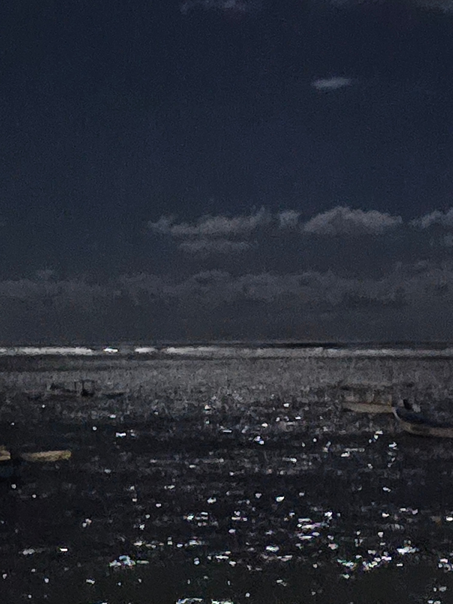 This is a dark, grainy, low-light image of a body of water, likely the sea, with a horizon line visible in the middle ground. The sky above is a deep, dark blue, with some scattered clouds. The water surface is dark and reflective, with numerous small, bright white specks, likely glints of light, scattered across it. In the water, there appear to be several indistinct shapes, some of which may be boats or buoys, with a few slightly lighter rectangular or elongated shapes visible near the horizon. The overall impression is of a nighttime scene, possibly during twilight or under a moonlit sky, given the darkness and the reflections on the water. The image does not contain any specific landmarks or details that would definitively place it in Denpasar, Indonesia.