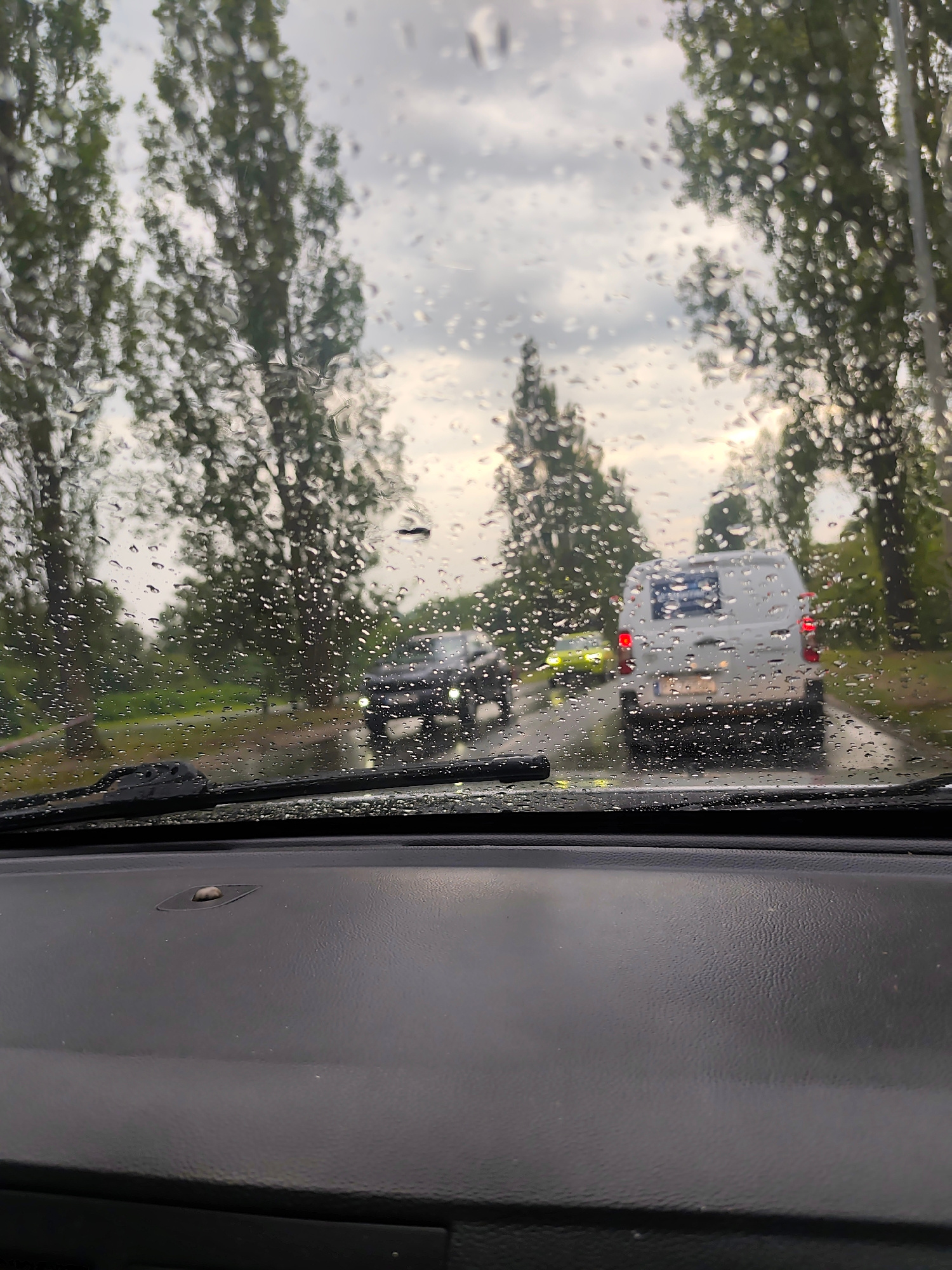 This media file captures a view from inside a vehicle, looking through a rain-streaked windshield. The scene depicts a road with several vehicles moving in the same direction. In the foreground, the dark dashboard of the car is visible, along with the windshield wipers. The road surface appears wet, indicating it is raining.
Beyond the windshield, tall trees with green foliage line the road on both sides, suggesting a suburban or semi-rural setting. The sky is overcast with clouds, typical of rainy weather. The time of day appears to be late afternoon or early evening, given the muted light.
The vehicles visible are a white van in the middle ground, a dark colored car to its left, and a bright yellow vehicle further ahead. The van has a blue and white sticker on its rear door. The license plate on the van is partially visible, but not legible.
The overall mood is one of driving in inclement weather. The rain drops on the windshield create a somewhat distorted and softened view of the outside world. The location context is identified as Szczecin, Poland.