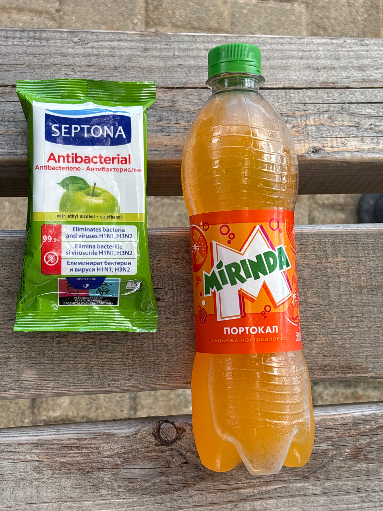 This is a top-down, eye-level, medium shot of two objects placed on a rustic wooden table. The scene is outdoors, as indicated by the weathered wood and the natural lighting suggesting daytime. The objects are a bottle of Mirinda orange soda and a pack of Septona antibacterial wipes.
The Mirinda bottle is to the right, partially filled with an orange-colored liquid. The label is predominantly orange and white, featuring the Mirinda logo and text in what appears to be Cyrillic and Latin alphabets. The text on the bottle includes "MIRINDA", "ПОРТОКАЛ" (Portokal, meaning Orange in Bulgarian), and "СЪДЪРЖА ПОРТОКАЛОВ СОК" (Contains orange juice). The bottle has a green cap.
Below the Mirinda bottle, to the left, is a green and white packet of Septona antibacterial wipes. The packet is sealed and displays text in multiple languages. Prominently visible are "SEPTONA" and "Antibacterial". Other text includes "with ethyl alcohol cu ethanol", "Eliminates bacteria and viruses H1N1, H3N2", and "99.9%". The packet also features an image of a green apple. There is a "PLASTIC IN PRODUCT" warning and instructions to "OPEN HERE". The time of day is not explicitly indicated, but the lighting suggests it is daylight. The setting is a weathered wooden surface, possibly a picnic table or outdoor seating area. No people are visible in the image. The overall mood is neutral and observational.