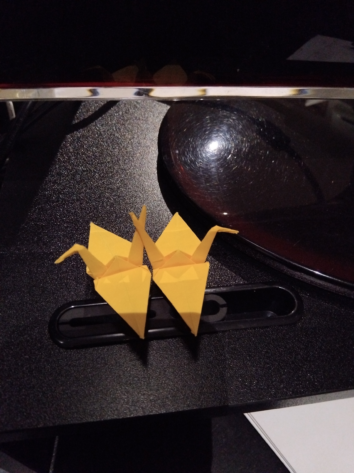 This image shows two yellow origami cranes placed on a textured black surface. The cranes are positioned side-by-side, with their beaks pointing slightly upwards and away from each other. They are resting on a black, elongated object with a cutout. In the background, a dark, glossy surface reflects light, creating a swirling pattern. The overall lighting suggests an indoor setting, possibly a desk or table. The image does not contain any information to determine a specific location in Vila Nova de Famalicão, Portugal.