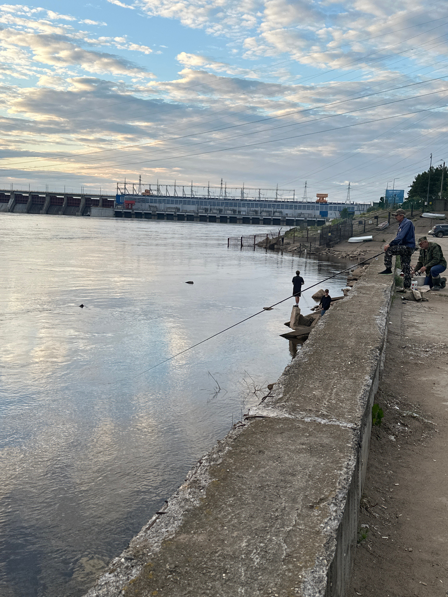 The image shows a wide river with a large dam structure in the background. Several people are fishing from the concrete embankment on the right side of the image. The sky is cloudy with patches of blue. The water reflects the sky and the dam. The embankment is rough and appears to be made of concrete. There are power lines visible in the sky above the dam.