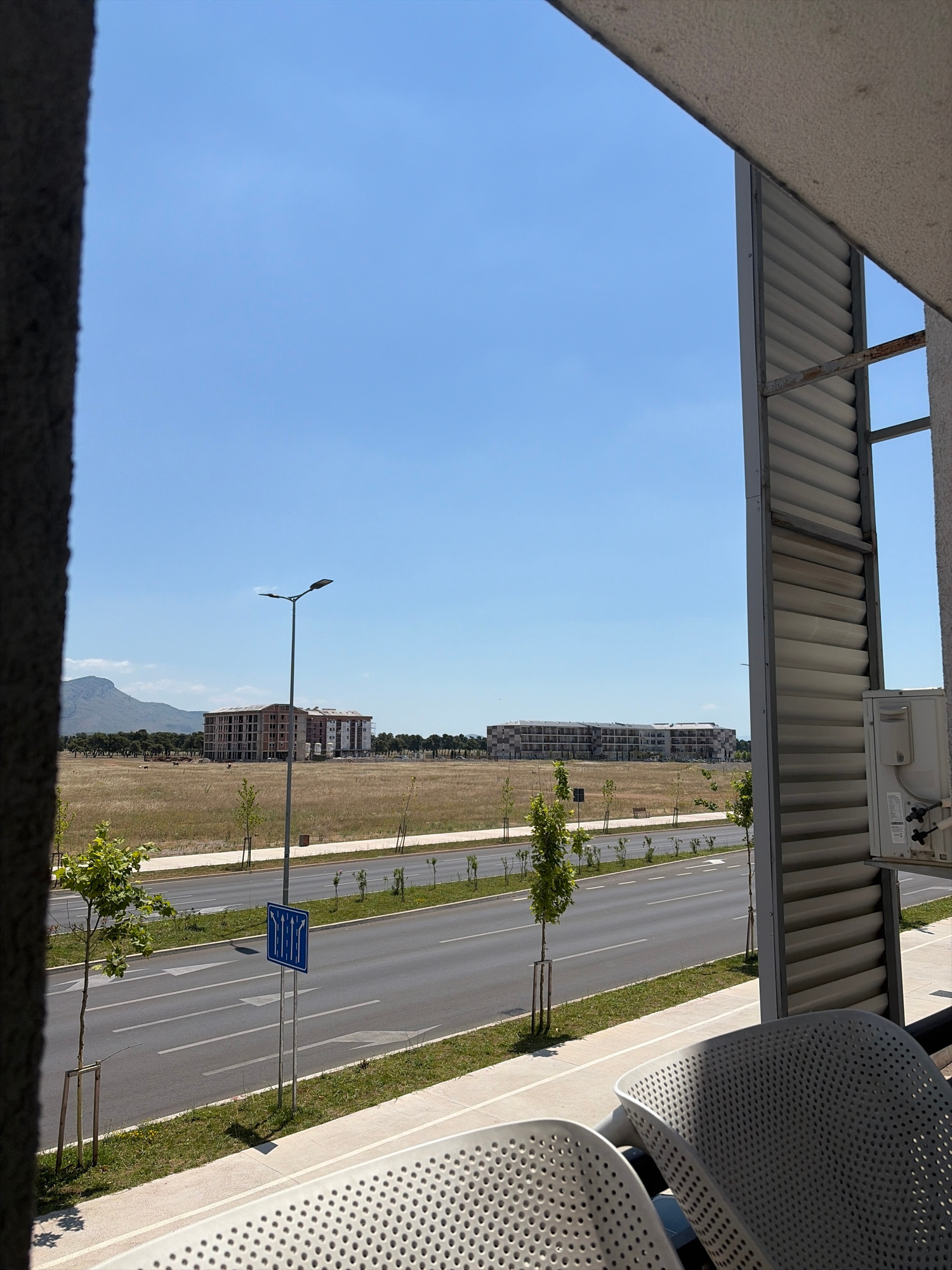 The image depicts a view from a balcony or elevated structure looking out over a street and an open field. The scene is set during the daytime, under a clear, bright blue sky, suggesting fair weather.
In the foreground, a portion of a concrete structure is visible, along with the textured surface of what appears to be furniture, possibly made of metal with a perforated design. Above, a corrugated metal roof structure with exposed metal beams is present.
The middle ground features a paved road with multiple lanes marked by white lines. Small trees are planted along the roadside, creating a lined effect. A blue traffic sign with white arrows indicating lane usage is prominently displayed next to the road. Beyond the road and grassy verge, a large field of dry, yellowish grass stretches out, dotted with more young trees.
In the background, several multi-story buildings, likely apartment complexes or offices, are under construction or recently completed, characterized by their unfinished facades and modular appearance. Further in the distance, a mountain range is visible against the horizon, partially obscured by a hazy atmosphere. The overall setting appears to be a developing urban or suburban area. No people are visible in the frame. The image is oriented vertically.