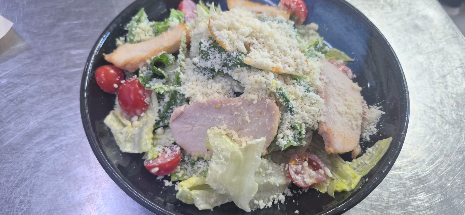 A close-up, overhead shot captures a serving of Caesar salad in a dark, circular bowl. The salad features ample shredded Parmesan cheese dusted over lettuce, sliced grilled chicken, and halved cherry tomatoes. The bowl rests on a metallic surface that appears to be a table or countertop, exhibiting a brushed texture with subtle scratches. The lighting is direct and somewhat harsh, creating visible reflections on the metallic surface. There are no people visible in the image, and no specific urban details that would indicate a location within Saint Petersburg, Russia.